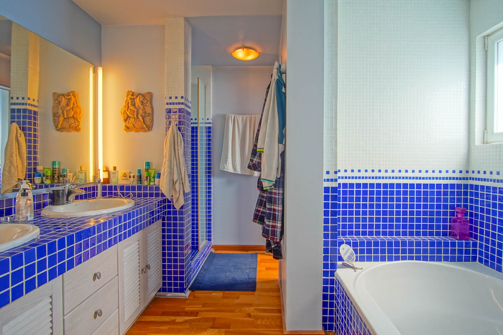 Baño con encimera de mosaico azul y blanco, lavabo doble, suelos de madera y una bañera integrada. Incluye iluminación cálida y armarios con puertas de lamas blancas.