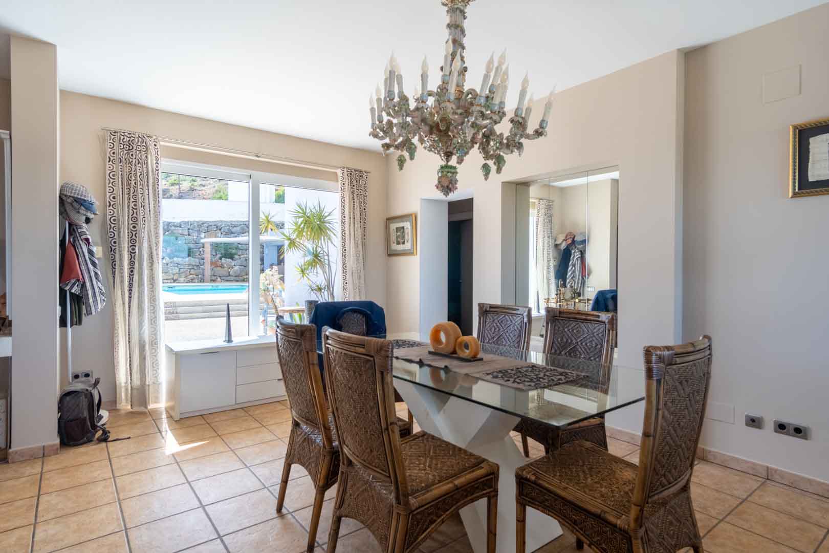 Comedor luminoso con mesa de cristal y sillas de mimbre, presidido por una lámpara de araña clásica y con vistas directas a la zona de la piscina a través de un gran ventanal.