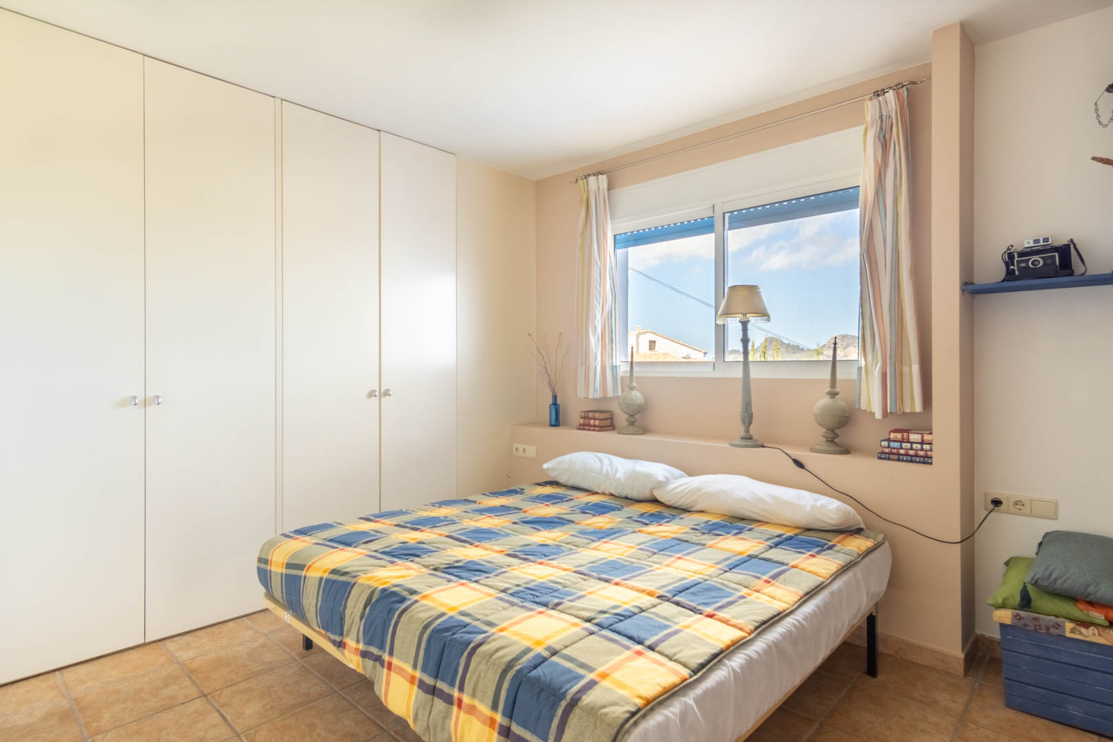 Dormitorio con amplios armarios empotrados blancos, suelos de gres rústico y una ventana que ofrece luz natural y vistas despejadas a las montañas.