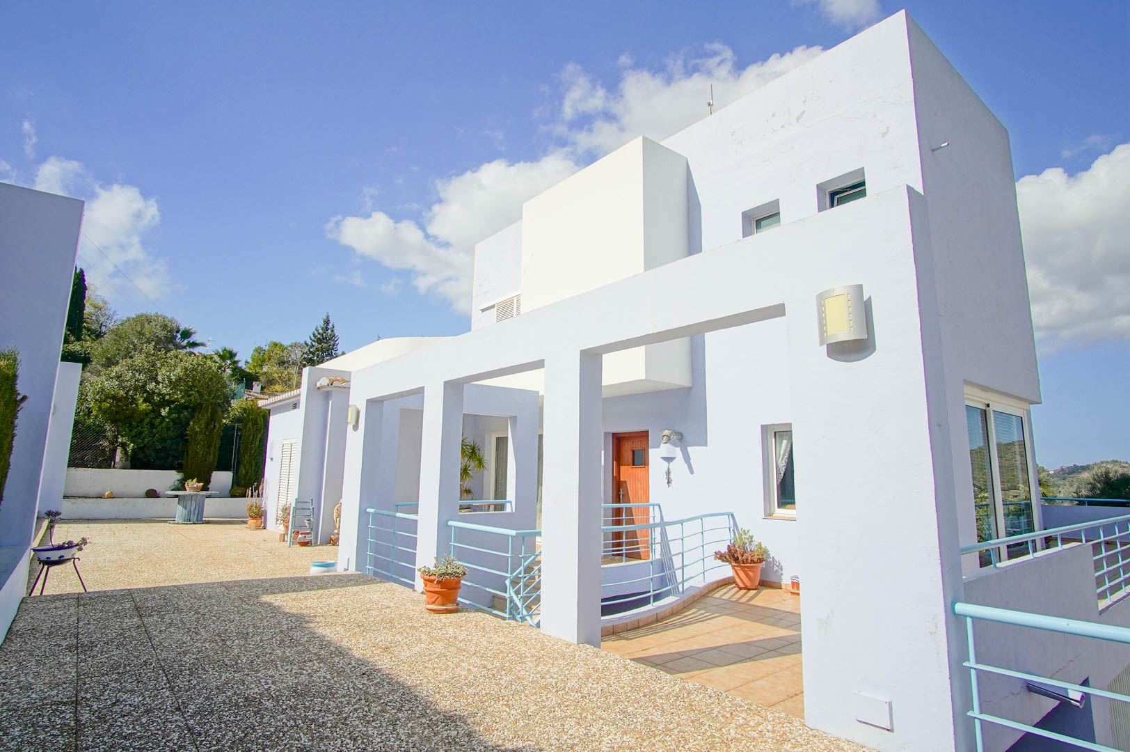 Fachada moderna de tonos claros con detalles arquitectónicos en azul, amplia terraza de grava y acceso principal con puerta de madera y barandillas metálicas.