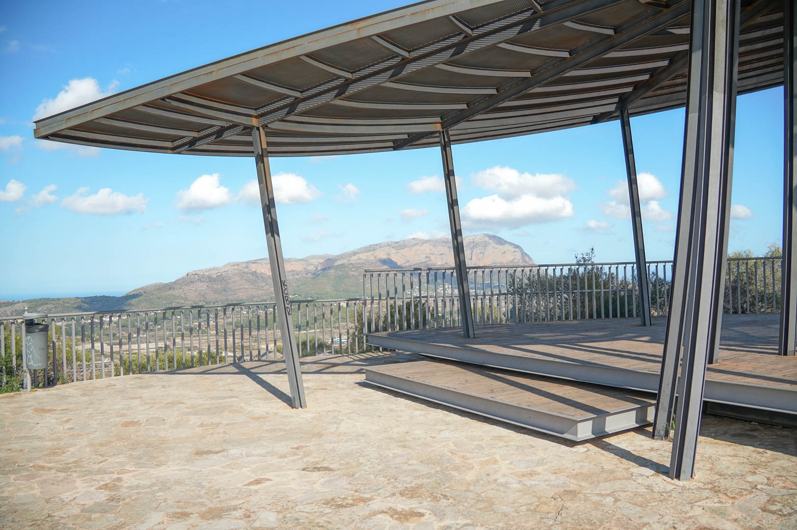 Mirador exterior con estructura metálica moderna y tarima de madera, ofreciendo vistas panorámicas a las montañas y al valle bajo un cielo despejado.