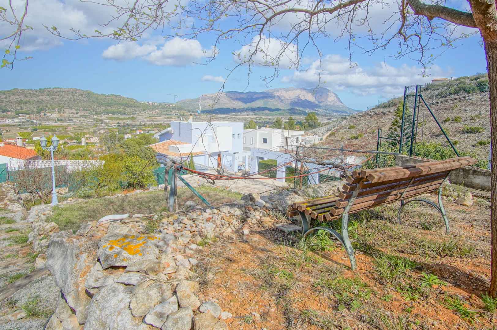 Mirador privado con banco de madera y murete de piedra natural, que ofrece vistas panorámicas despejadas hacia el valle y las montañas bajo el cielo mediterráneo.
