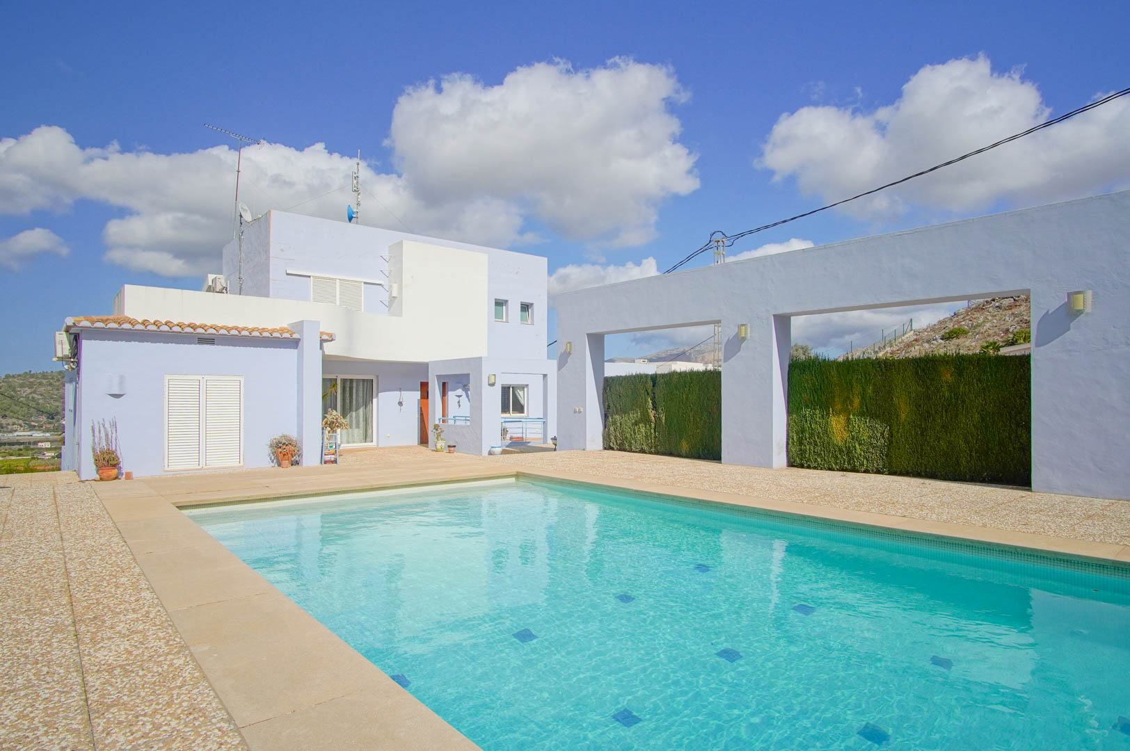 Piscina rectangular rodeada de una amplia terraza de piedra, con la fachada moderna de tonos azulados y blancos de la villa al fondo bajo un cielo despejado.