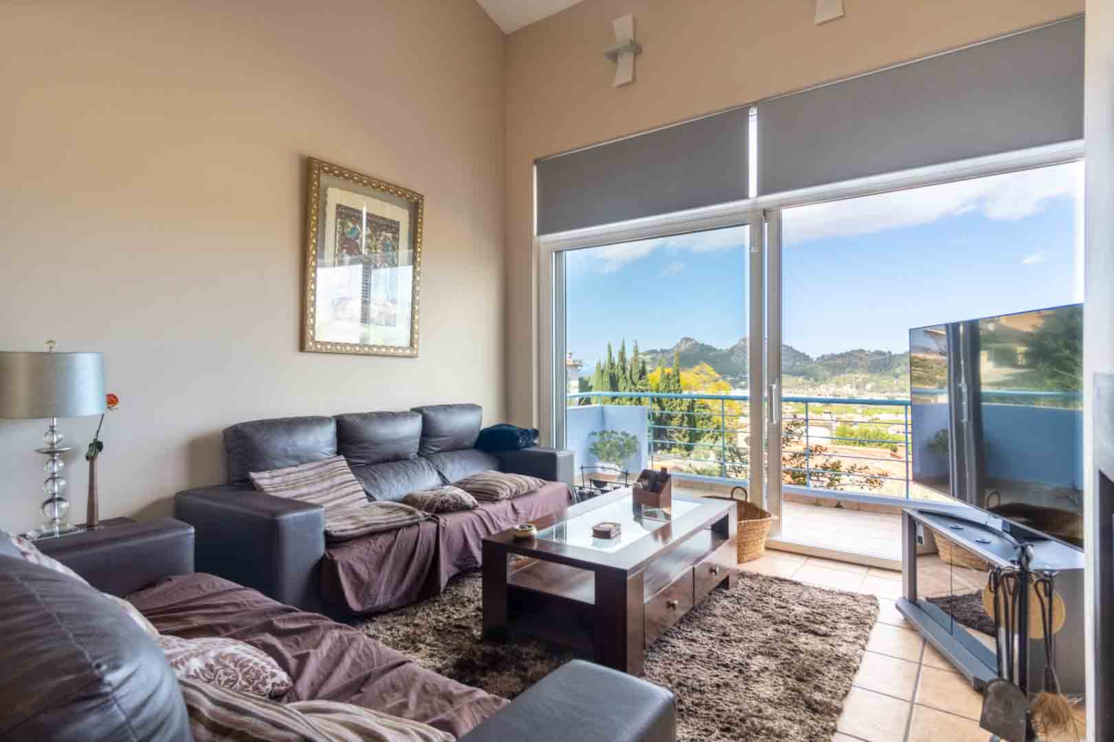 Salón con techos altos, sofás oscuros y mesa de centro de madera. Un gran ventanal da acceso a la terraza con vistas panorámicas a las montañas de Pedreguer.