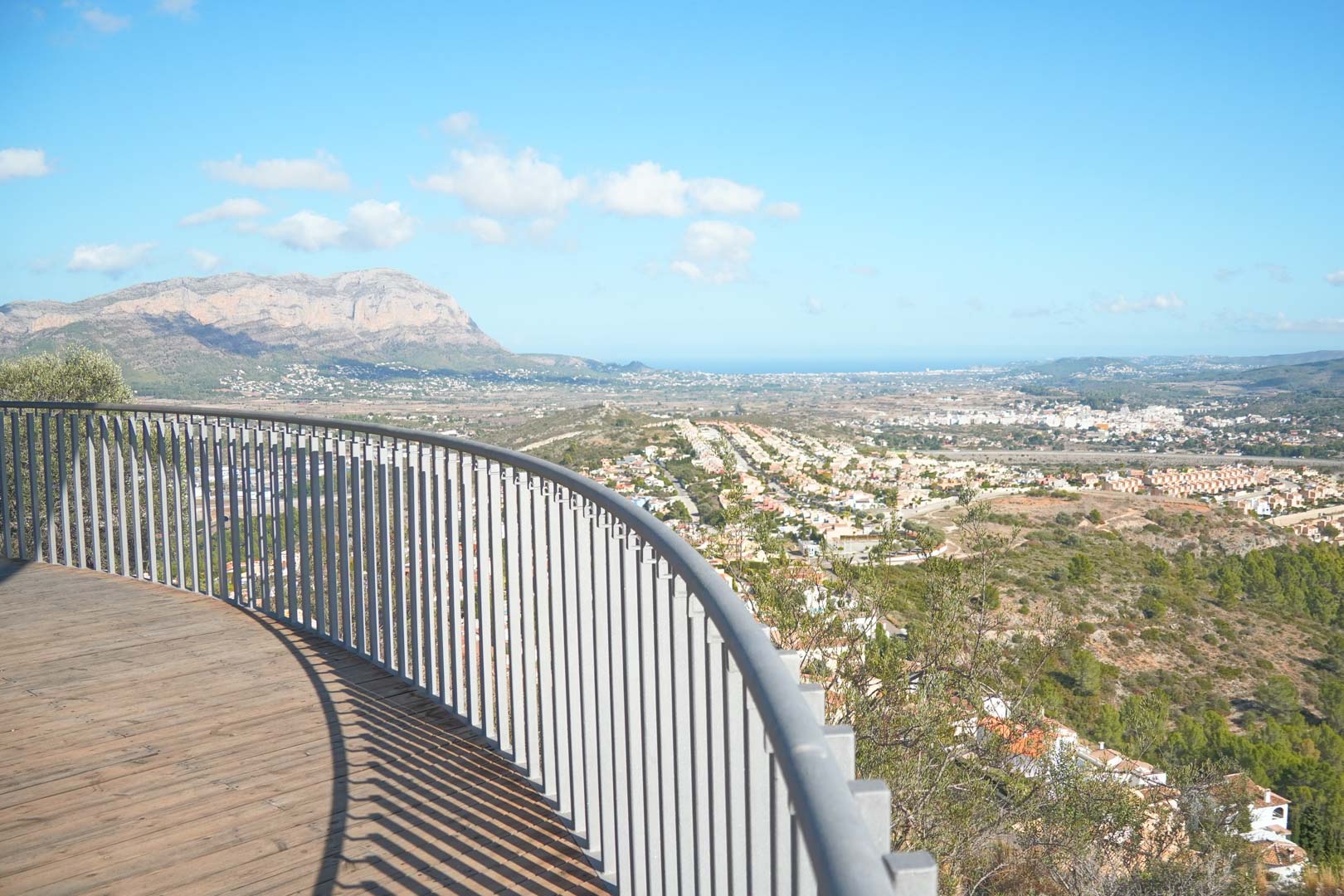 Terraza curva con suelo de madera y barandilla metálica que ofrece vistas panorámicas al valle, las montañas y el mar Mediterráneo bajo un cielo despejado.