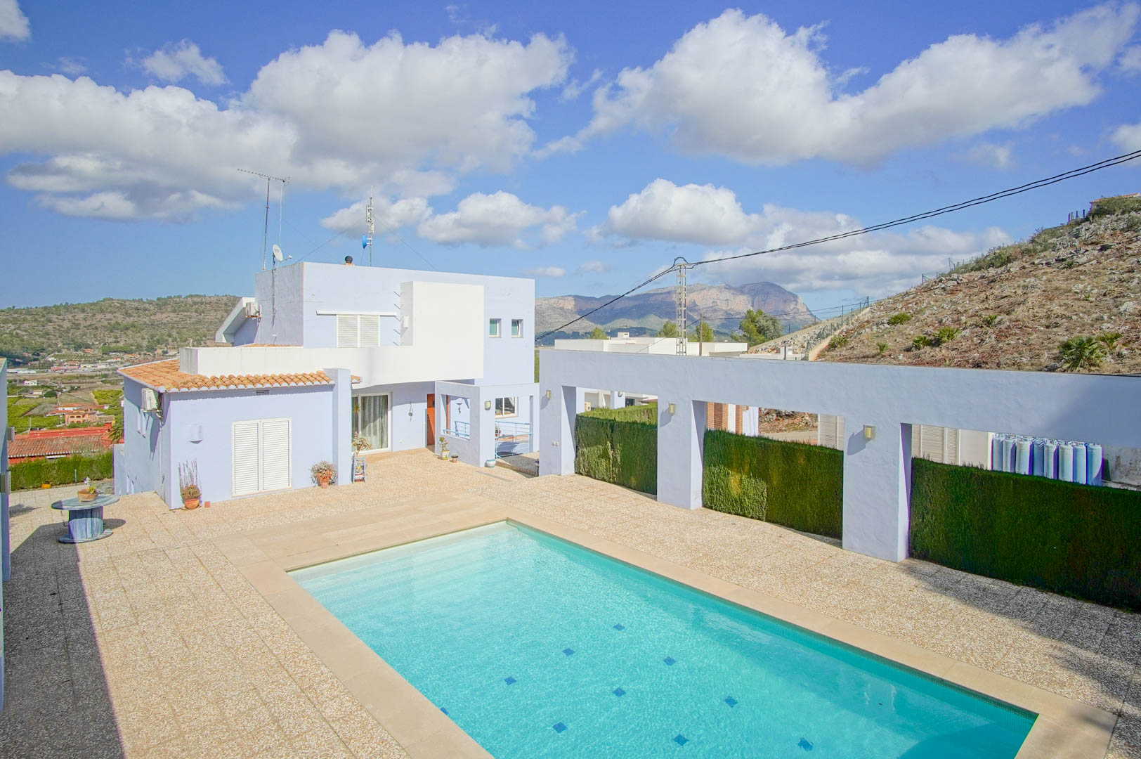 Vista de la piscina rectangular y terraza de piedra con la fachada lavanda de la villa y montañas al fondo bajo un cielo despejado.