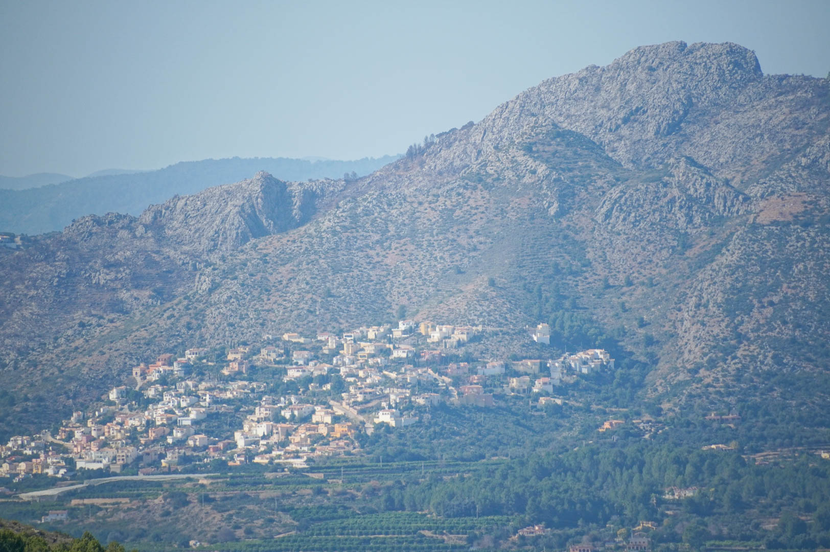 Vista panorámica de las montañas y el valle, con una urbanización residencial integrada en la ladera y campos agrícolas mediterráneos en la base.