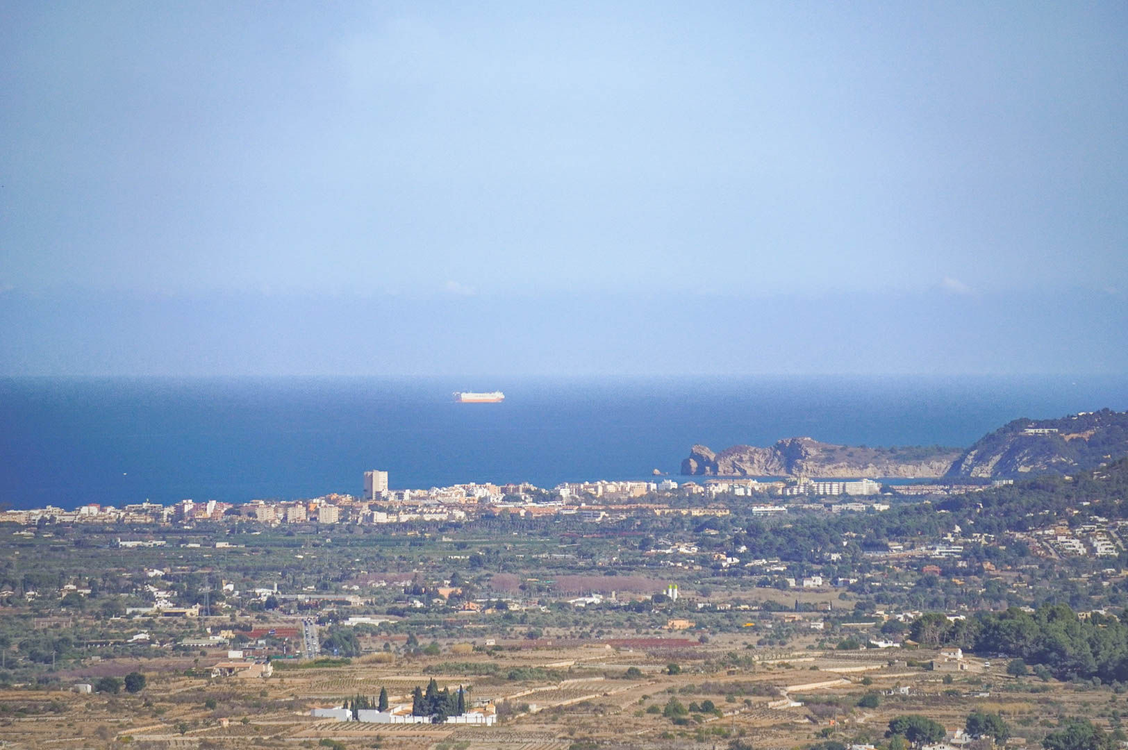 Vistas panorámicas despejadas hacia la costa mediterránea, mostrando el perfil urbano de la ciudad costera y el mar con embarcaciones en el horizonte.