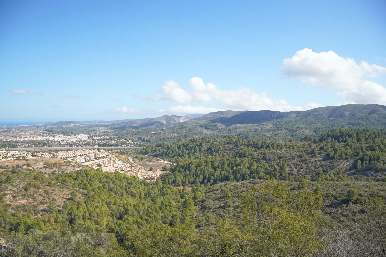 Vistas panorámicas hacia el valle de Pedreguer, mostrando montañas cubiertas de pinos, núcleos urbanos y el mar Mediterráneo en el horizonte bajo un cielo despejado.