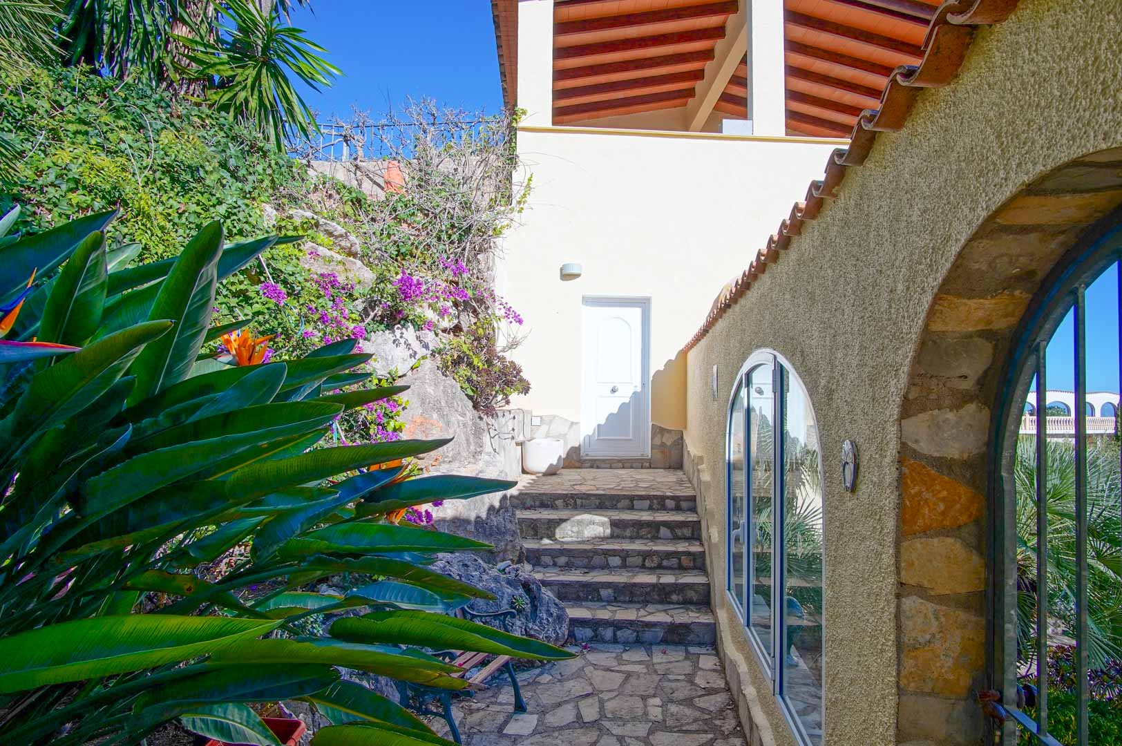 Acceso exterior con escaleras de piedra natural, rodeado de vegetación mediterránea y muros de piedra rústica con ventanales de arco.