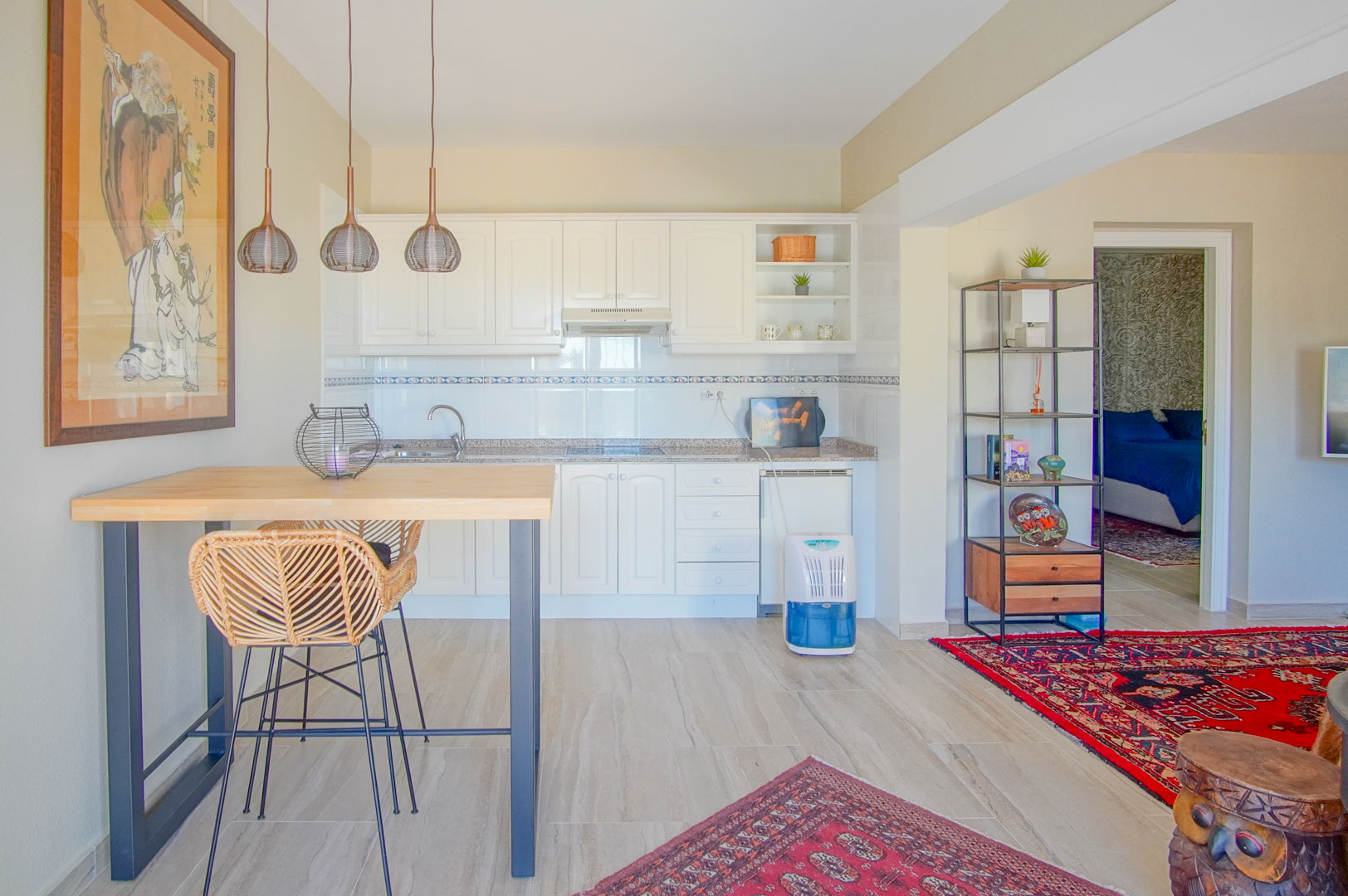 Cocina abierta con armarios blancos, encimera de granito y barra de madera con taburete de mimbre. El espacio conecta con una zona de estar decorada con alfombras orientales.