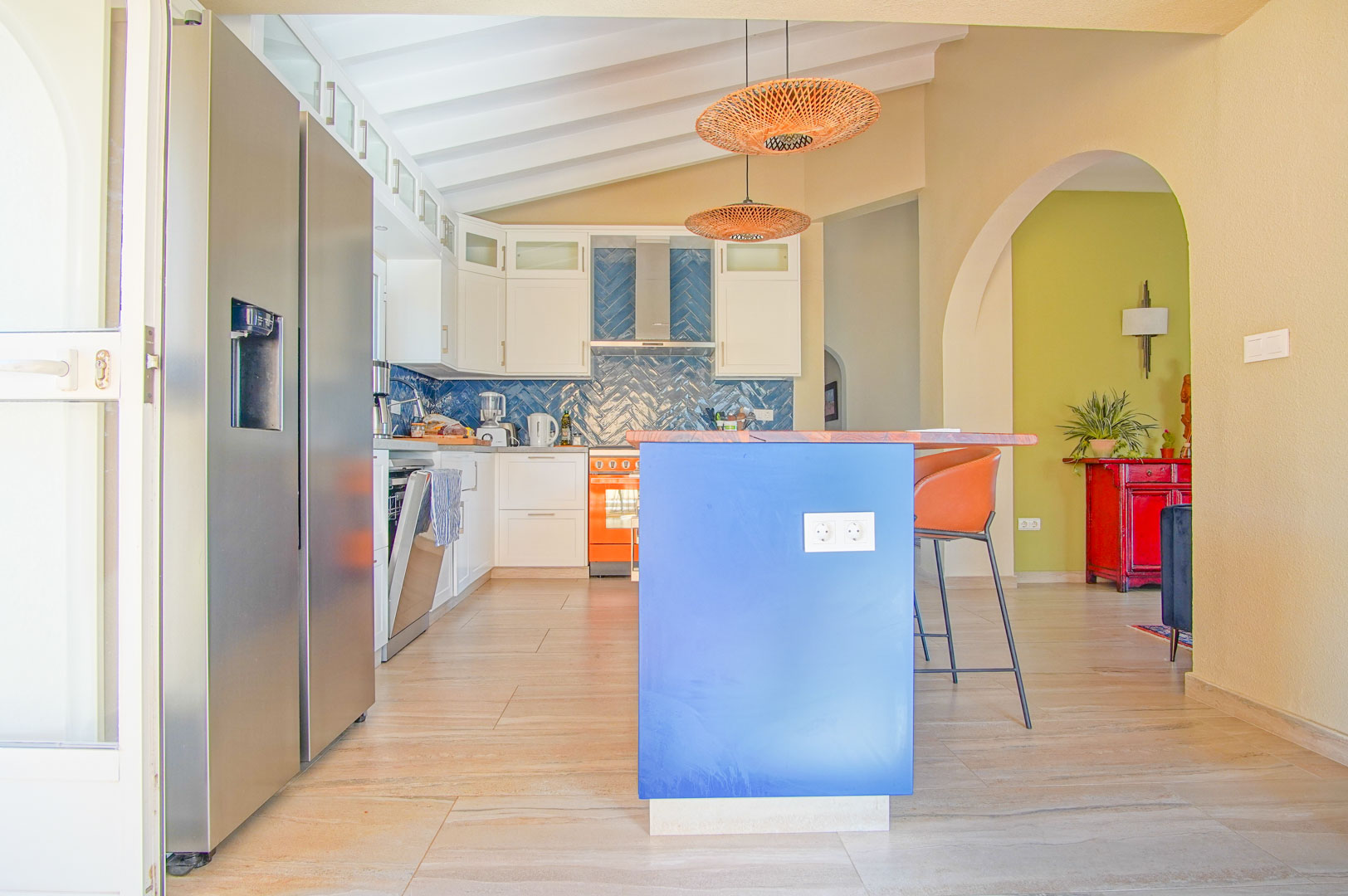 Cocina equipada con isla azul, encimera de madera y salpicadero de azulejos en espiga. Destacan los techos de vigas blancas, lámparas de fibras naturales y electrodomésticos modernos.