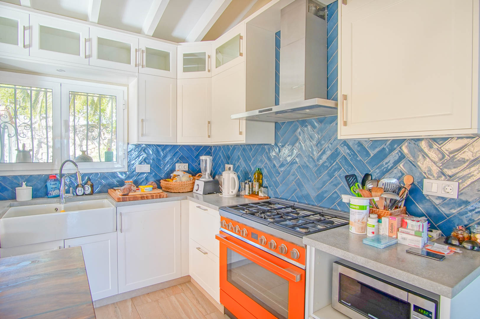 Cocina luminosa con azulejos azules en espiga, encimera gris y un horno naranja distintivo. Incluye fregadero cerámico y vigas de madera blancas en el techo.