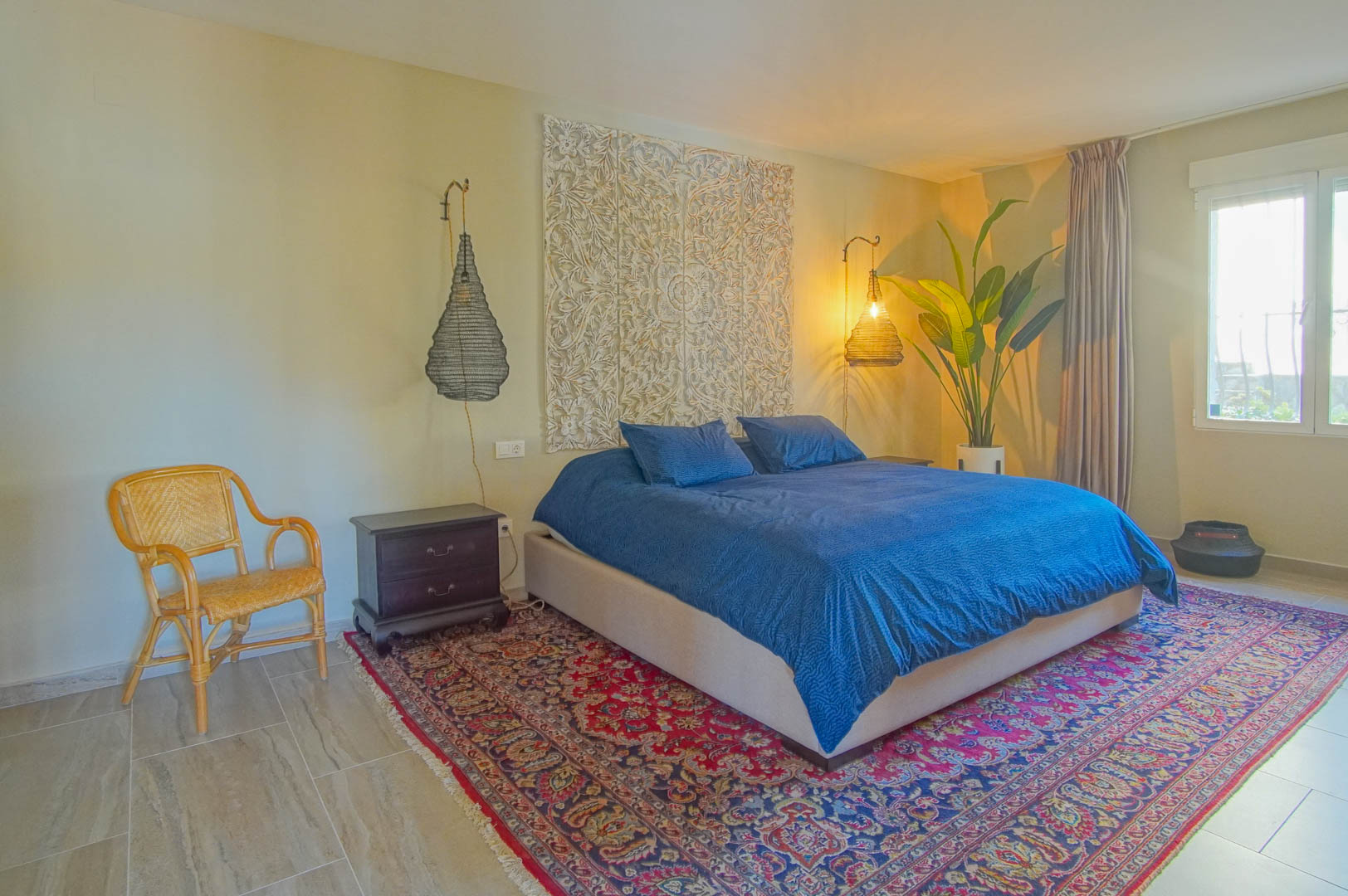 Dormitorio con cabecero de madera tallada, lámparas colgantes de mimbre y suelos cerámicos. Incluye una alfombra de estilo oriental y acceso a luz natural.