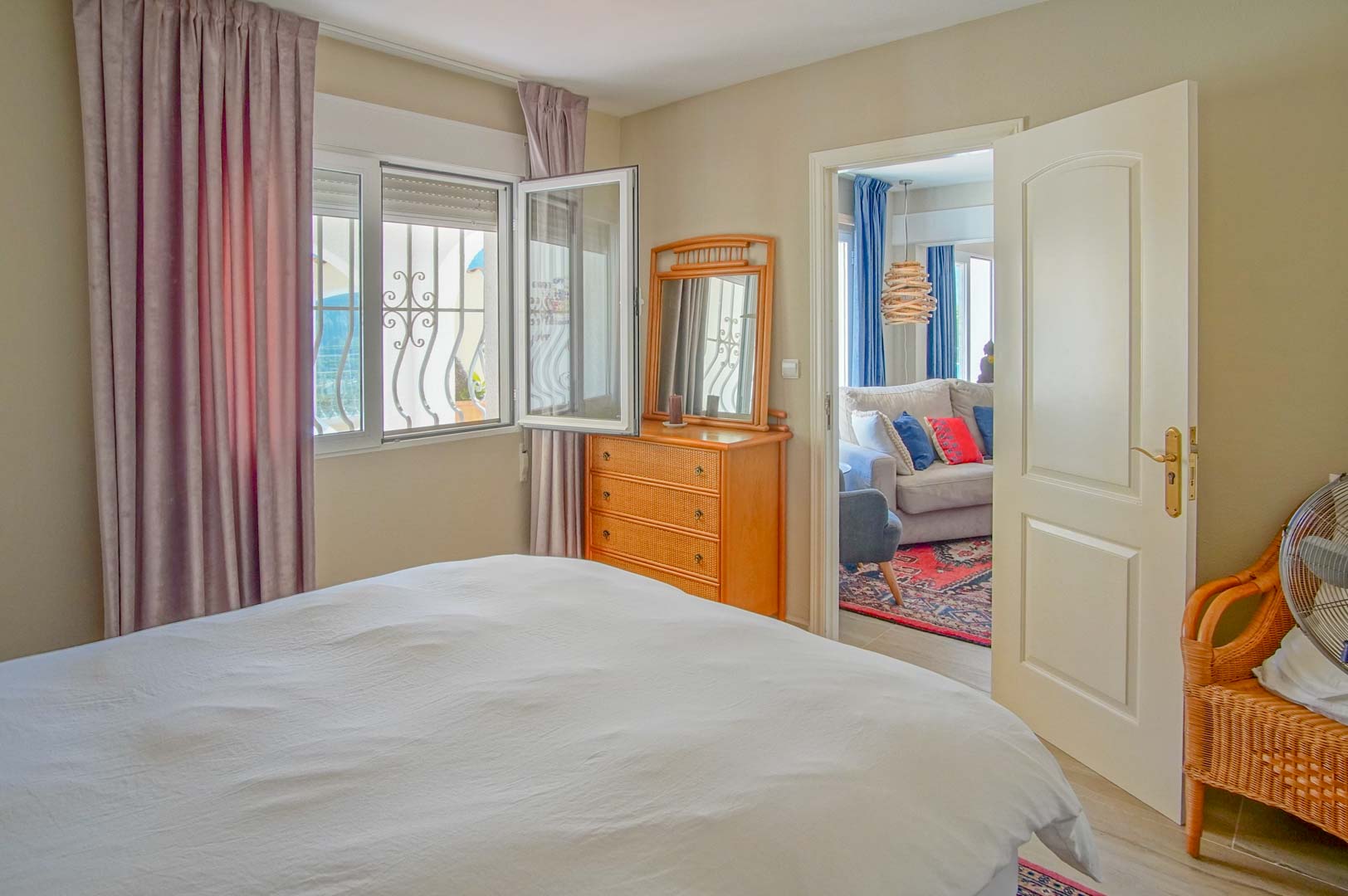 Dormitorio luminoso con cómoda de madera y ventana con rejas decorativas. La puerta abierta revela un salón contiguo con sofá y alfombra de estilo tradicional.
