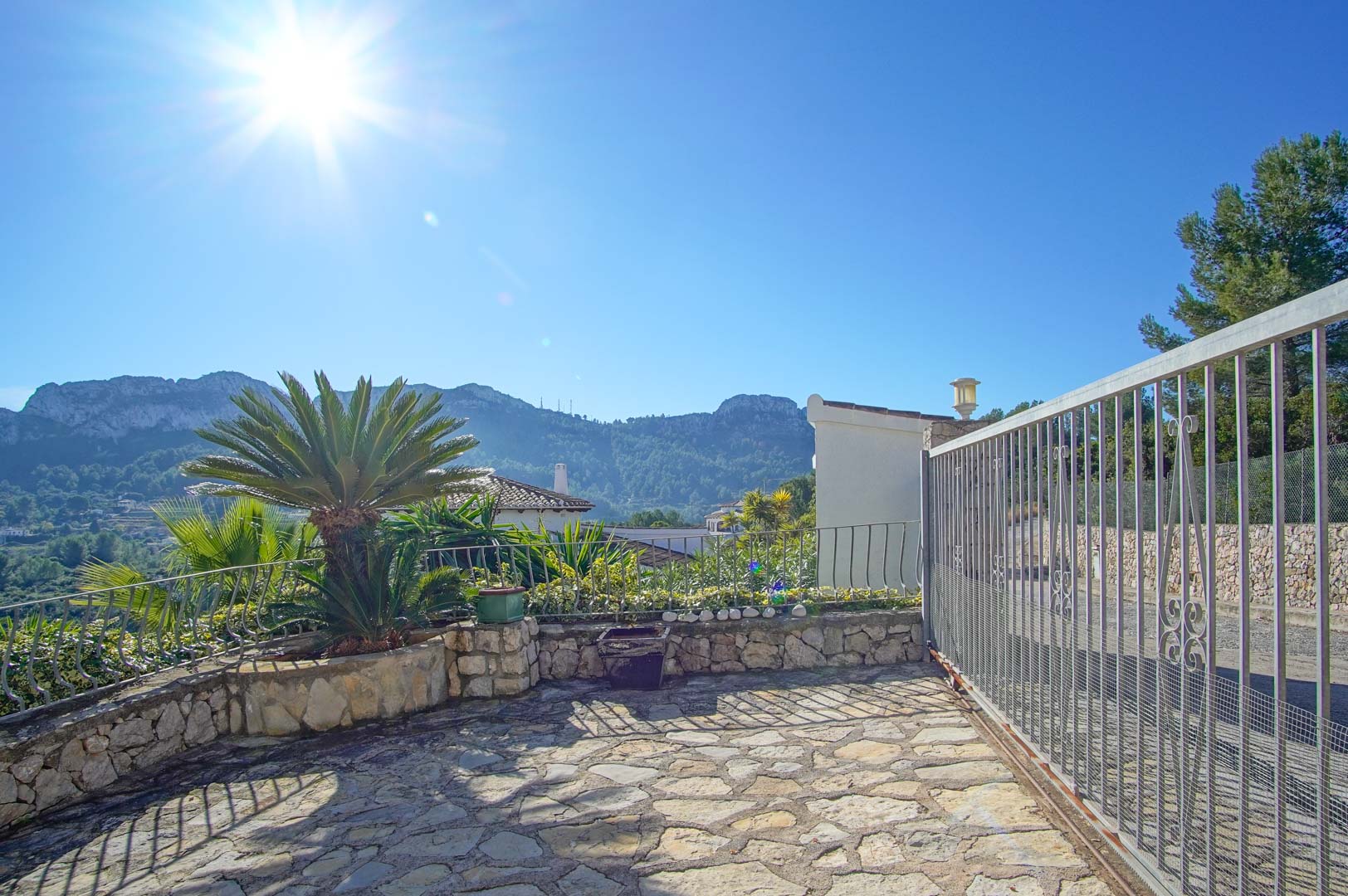 Entrada a la propiedad con suelo de piedra natural, puerta corredera metálica y vistas despejadas a las montañas de Monte Pego bajo un cielo despejado.