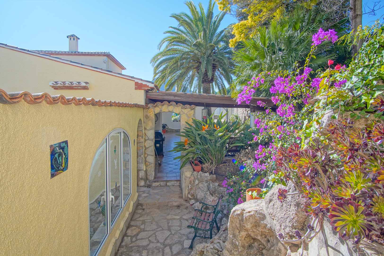 Entrada exterior con pavimentación de piedra natural, rodeada de buganvillas y palmeras, que conduce a un porche cubierto mediante un arco de piedra rústica.