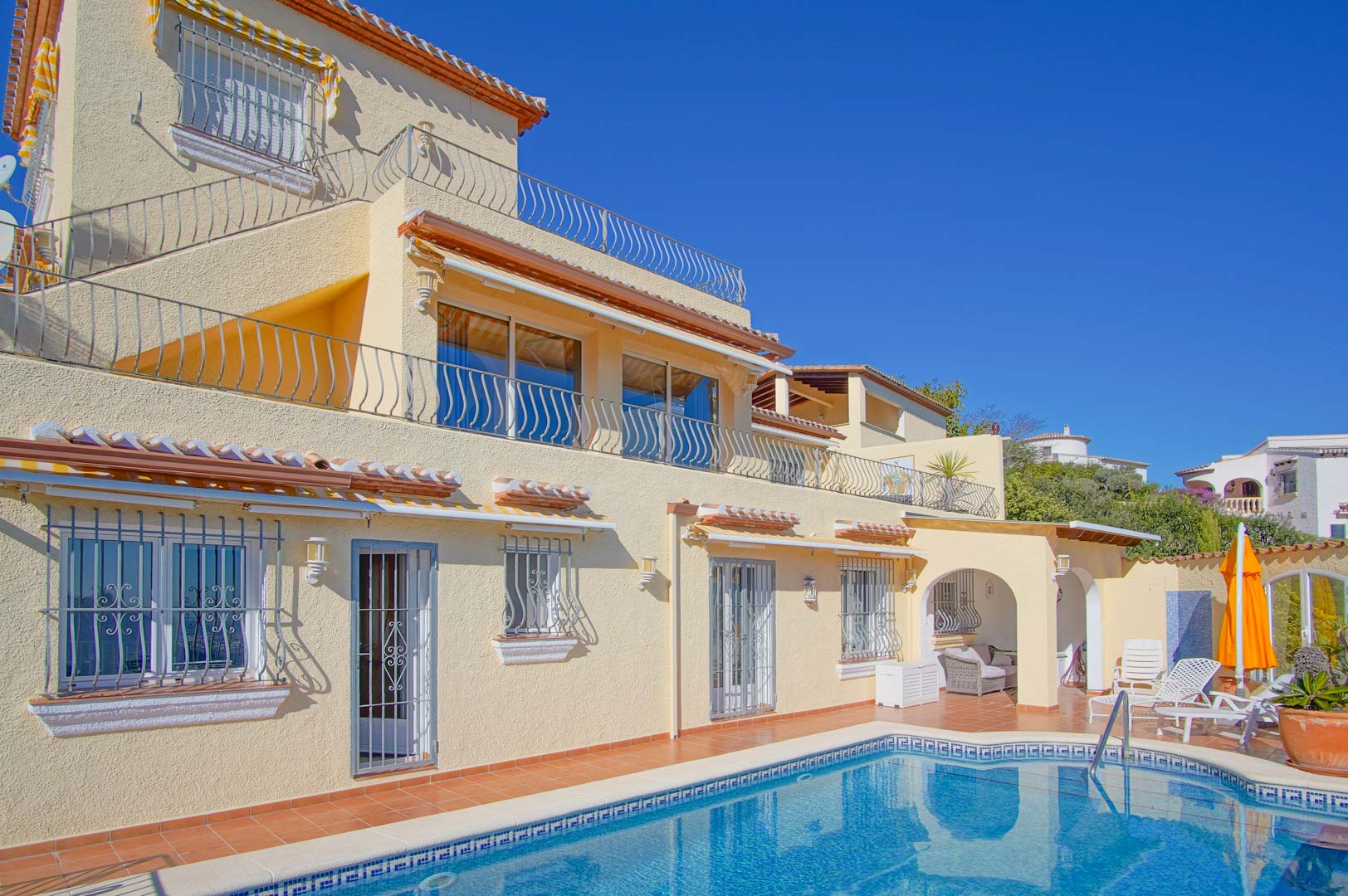 Fachada de tres niveles con piscina de mosaico azul, amplias terrazas con barandillas de forja y zona de porche cubierto junto al solárium de terracota.