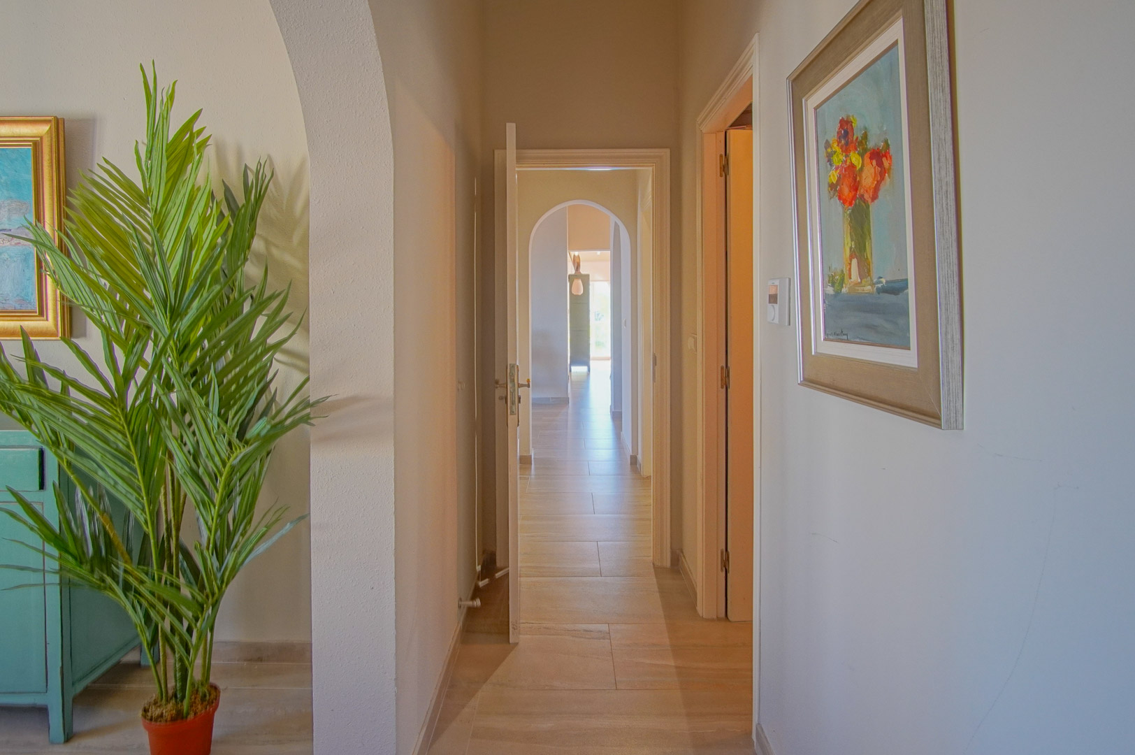 Pasillo interior con suelos cerámicos, arcos decorativos y acceso a diversas estancias. La iluminación natural resalta las paredes neutras y la decoración artística.