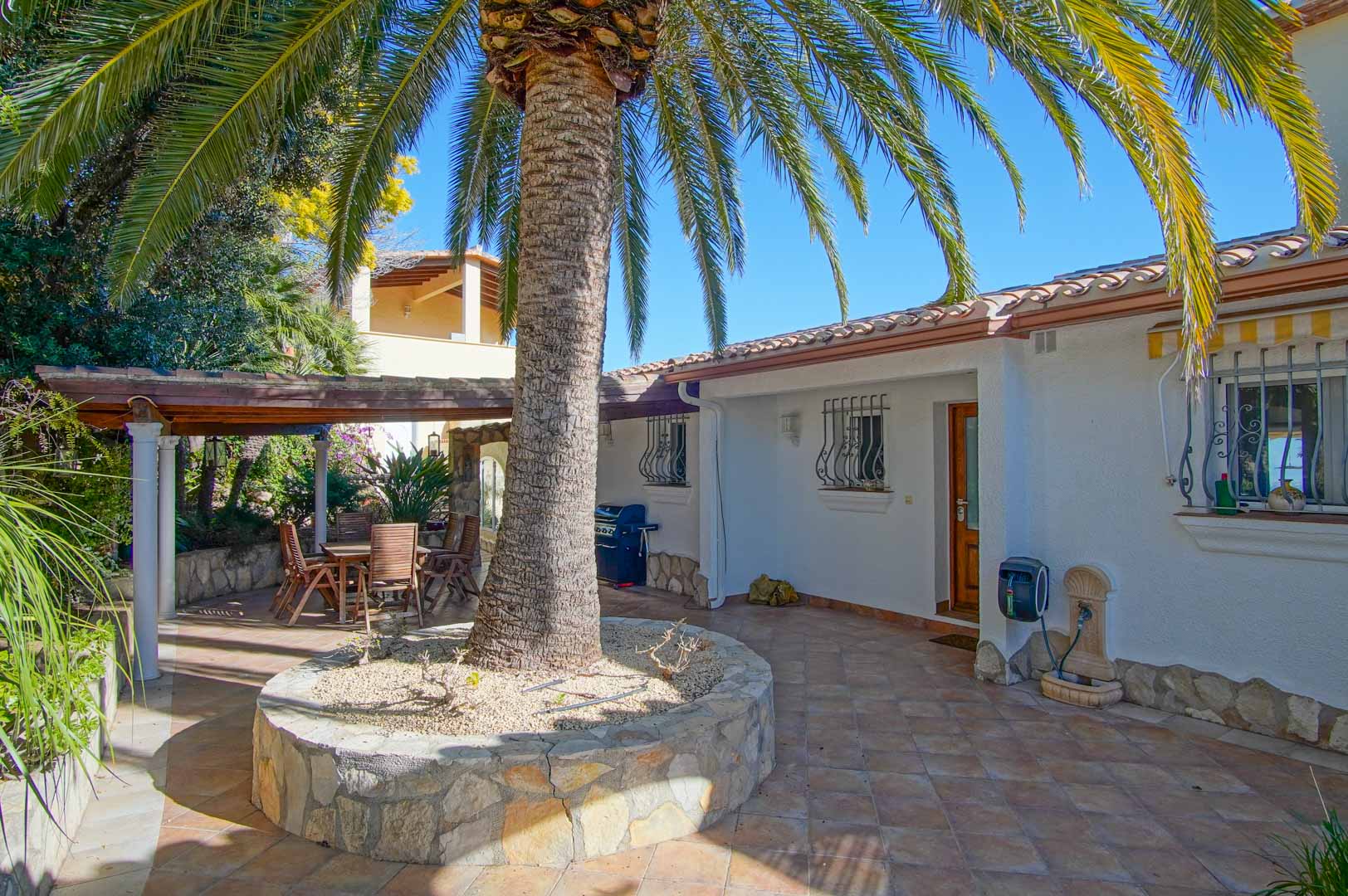 Patio exterior con suelo cerámico, una palmera central en jardinera de piedra y zona de comedor bajo pérgola de madera rodeada de vegetación mediterránea.