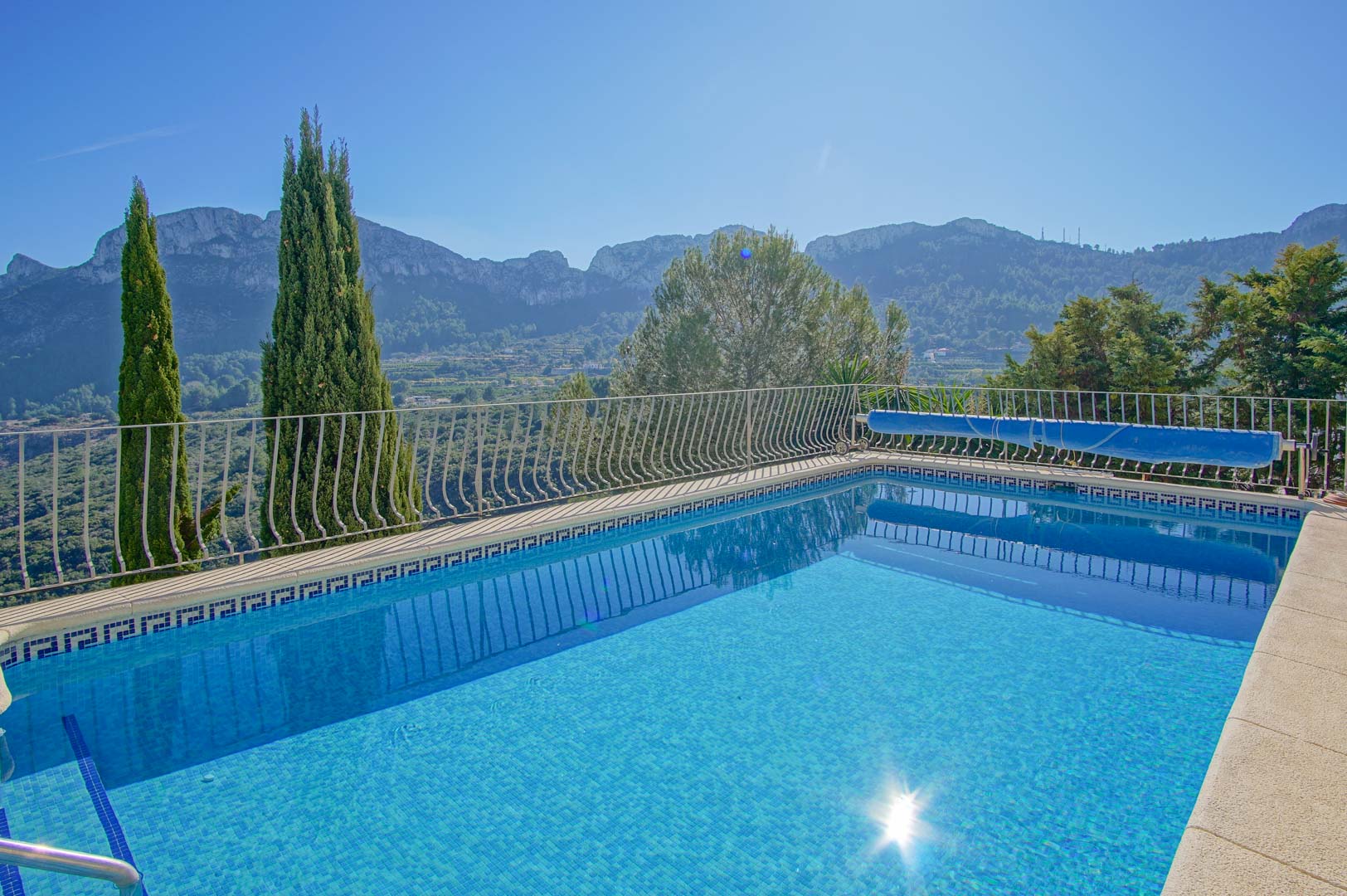 Piscina exterior con revestimiento de gresite azul, barandilla de hierro y vistas panorámicas a las montañas y al valle de Monte Pego bajo un cielo despejado.
