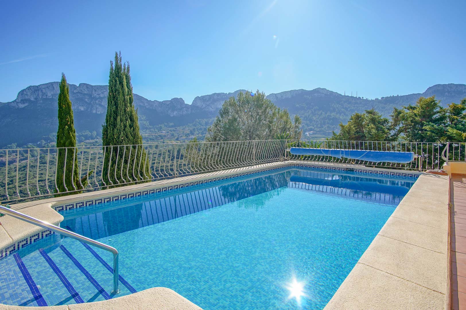Piscina exterior con revestimiento de gresite azul, escalera de obra y barandilla metálica, ofreciendo vistas despejadas a las montañas de Monte Pego.