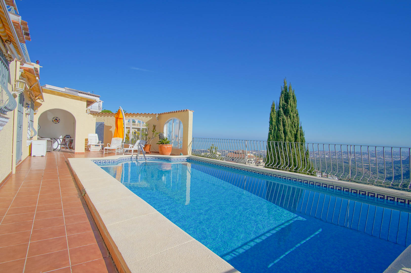 Piscina rectangular con mosaico azul y cenefa griega, terraza de terracota y vistas despejadas al mar Mediterráneo y al valle de Monte Pego.