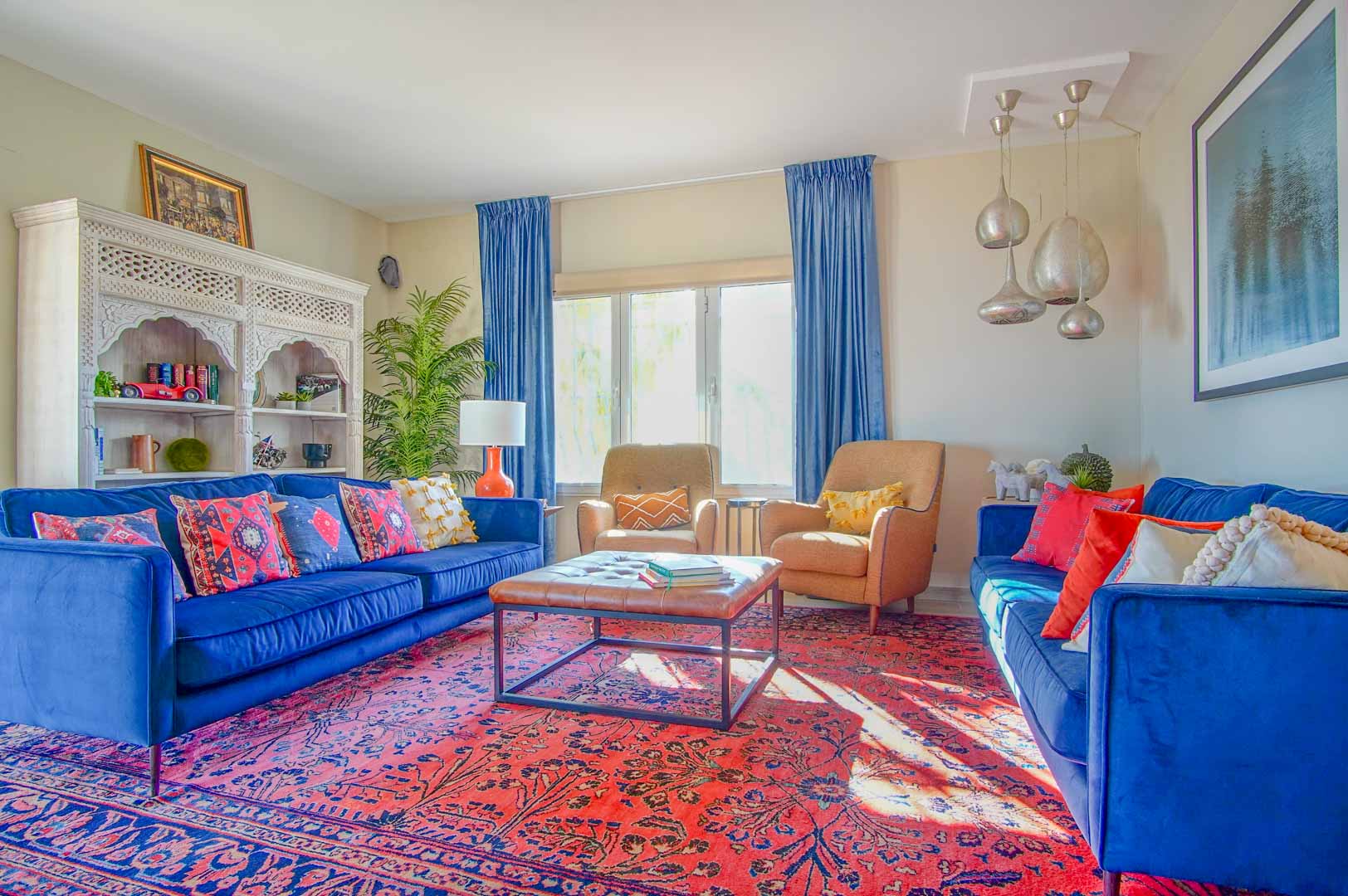 Salón con sofás de terciopelo azul, alfombra oriental roja y estantería de madera tallada. La estancia cuenta con iluminación natural y lámparas colgantes plateadas.