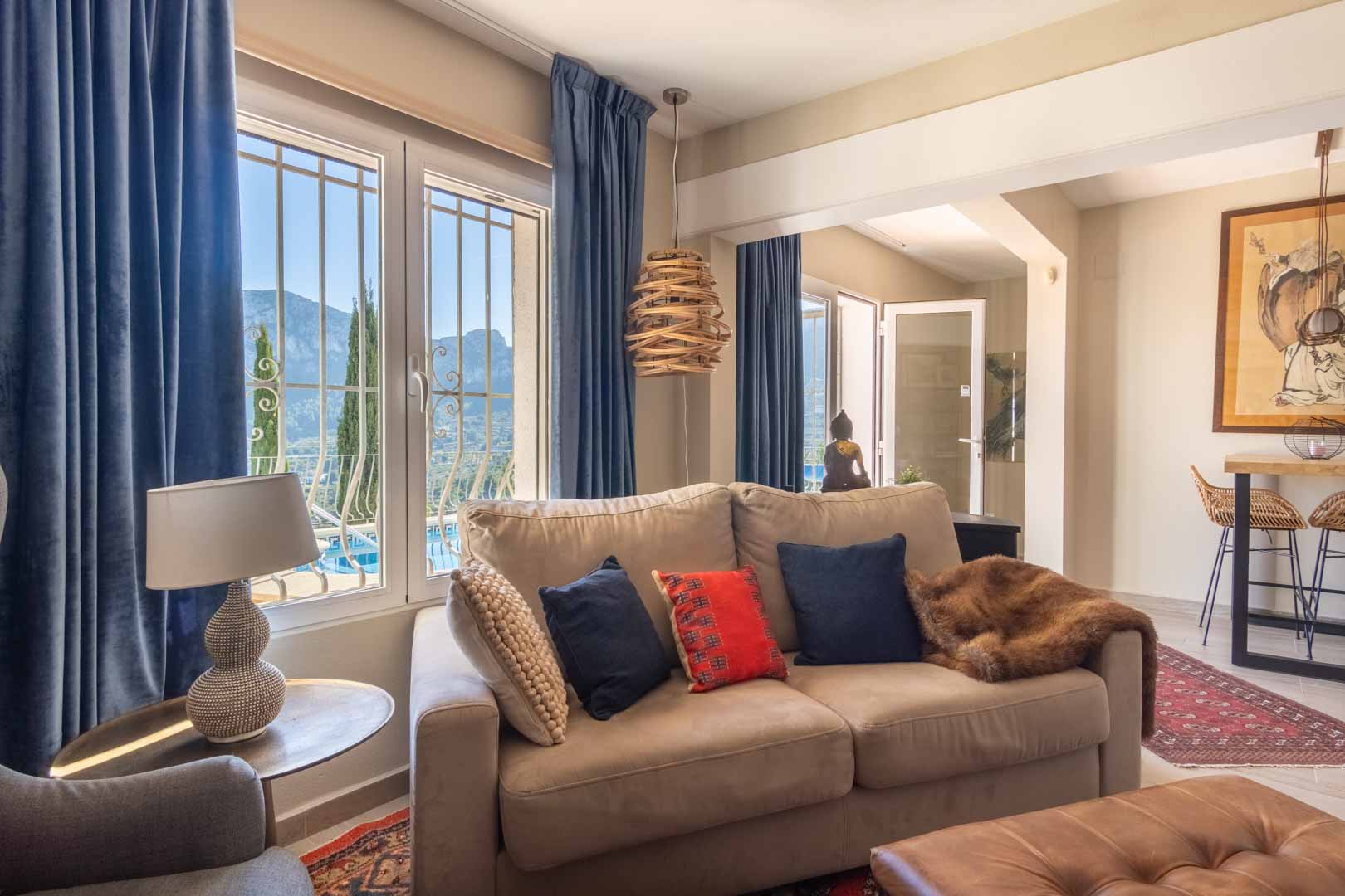 Salón luminoso con sofá en tonos tierra, cortinas de terciopelo azul y ventanales con vistas a la montaña. Incluye una zona de barra contigua y decoración de estilo étnico.