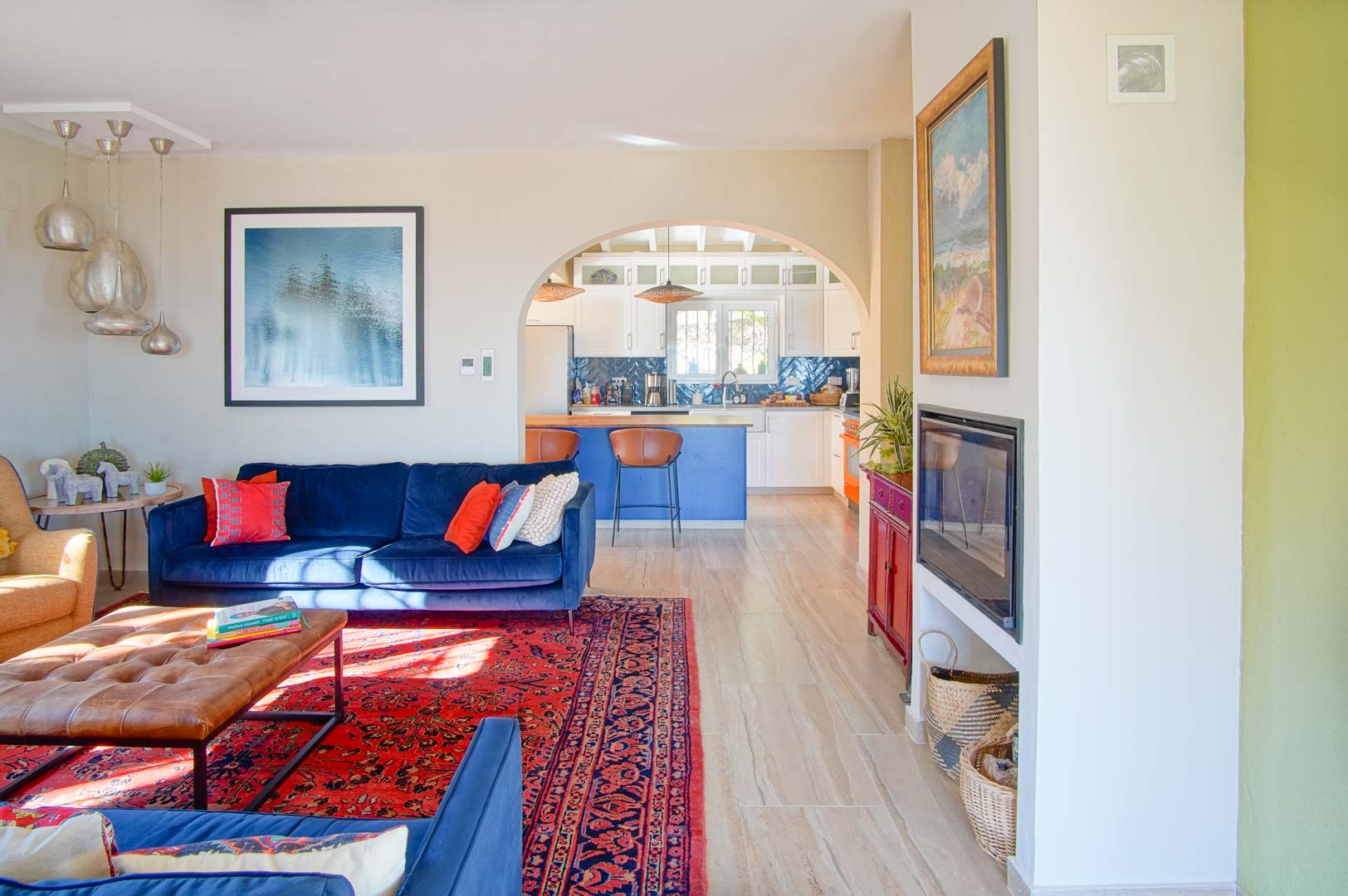 Salón luminoso con sofás de terciopelo azul, chimenea encastrada y alfombra oriental, conectado mediante un arco a una cocina con isla azul y encimera de madera.