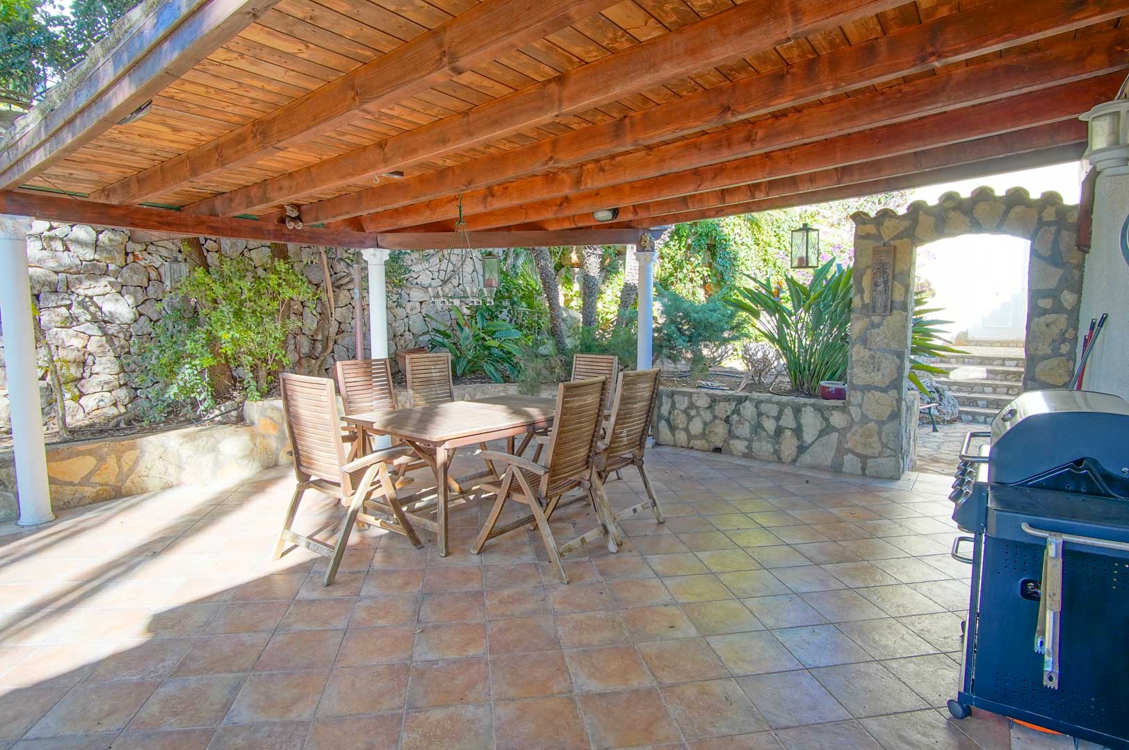 Terraza cubierta con vigas de madera que alberga un comedor exterior, barbacoa y muros decorativos de piedra natural integrados en el jardín.
