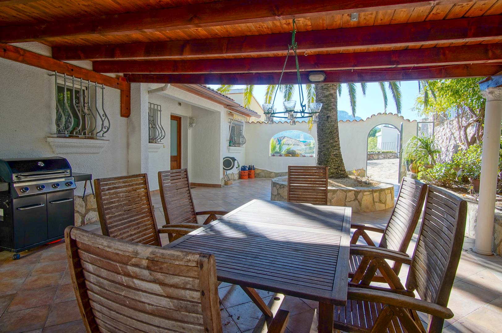 Terraza cubierta con vigas de madera, zona de comedor exterior, barbacoa de gas y patio con palmera rodeada de muros de piedra natural.