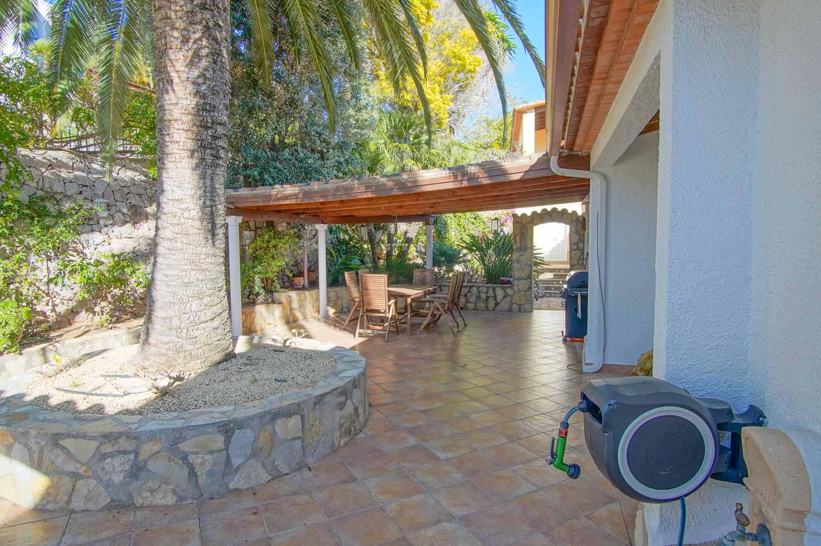 Terraza exterior con suelo cerámico, zona de comedor bajo pérgola de madera y muros de piedra natural integrados con la vegetación mediterránea.