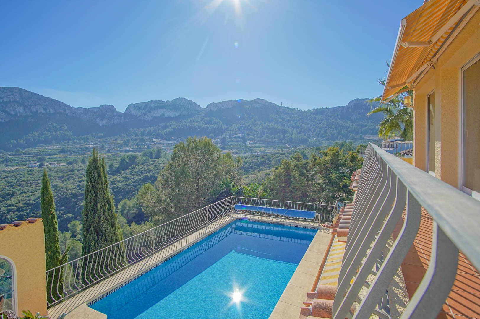 Vista elevada de la piscina rectangular con barandilla metálica, rodeada de vegetación y vistas panorámicas a las montañas de la Marina Alta.