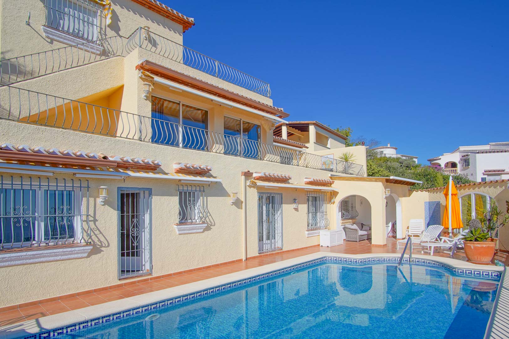 Vista exterior de la villa con piscina privada, terrazas de terracota y fachada de tres niveles. Incluye zona de chill-out bajo arco y barandillas de hierro forjado.