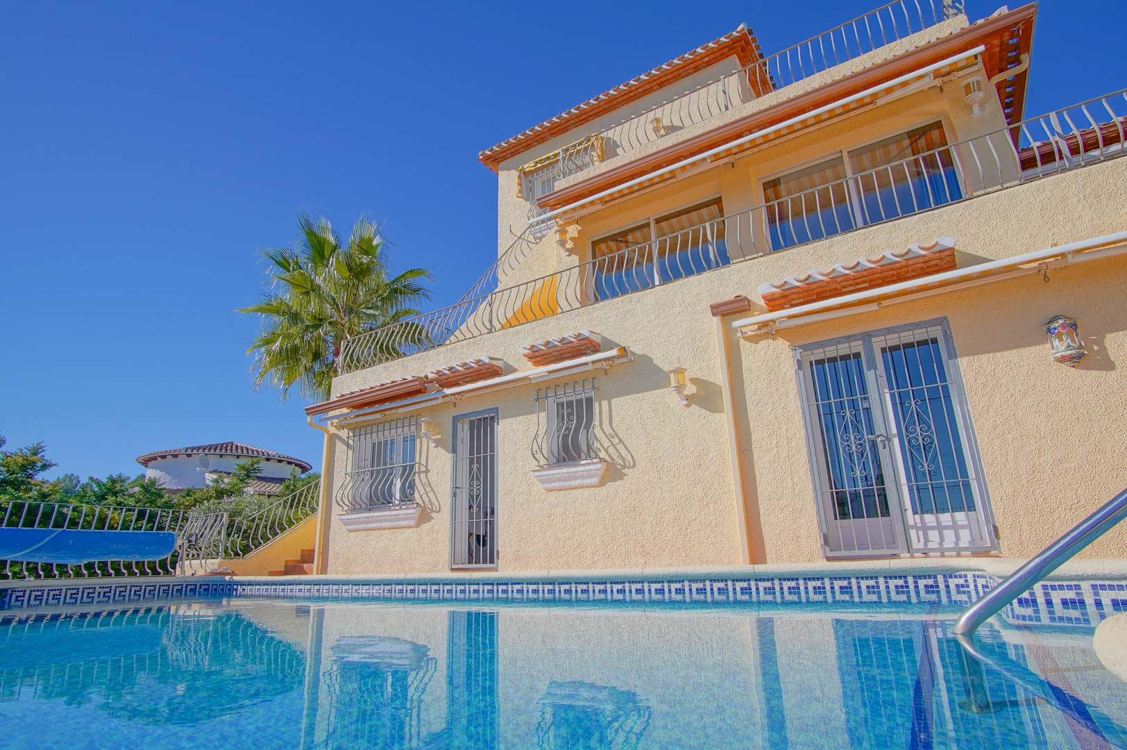 Vista exterior de la villa de tres niveles desde la piscina, con terrazas escalonadas, barandillas de hierro forjado y fachada en tonos crema.