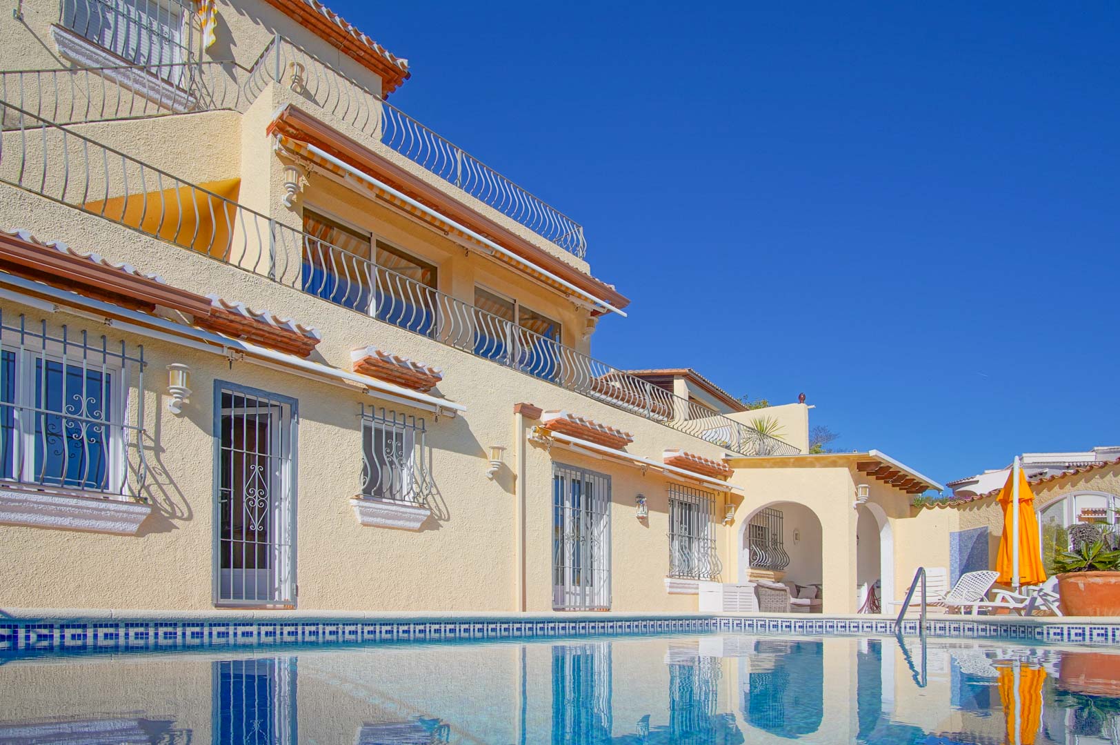 Vista exterior desde la piscina que muestra la fachada de tres niveles con terrazas, barandillas de hierro forjado y zona de chill-out junto al solárium.
