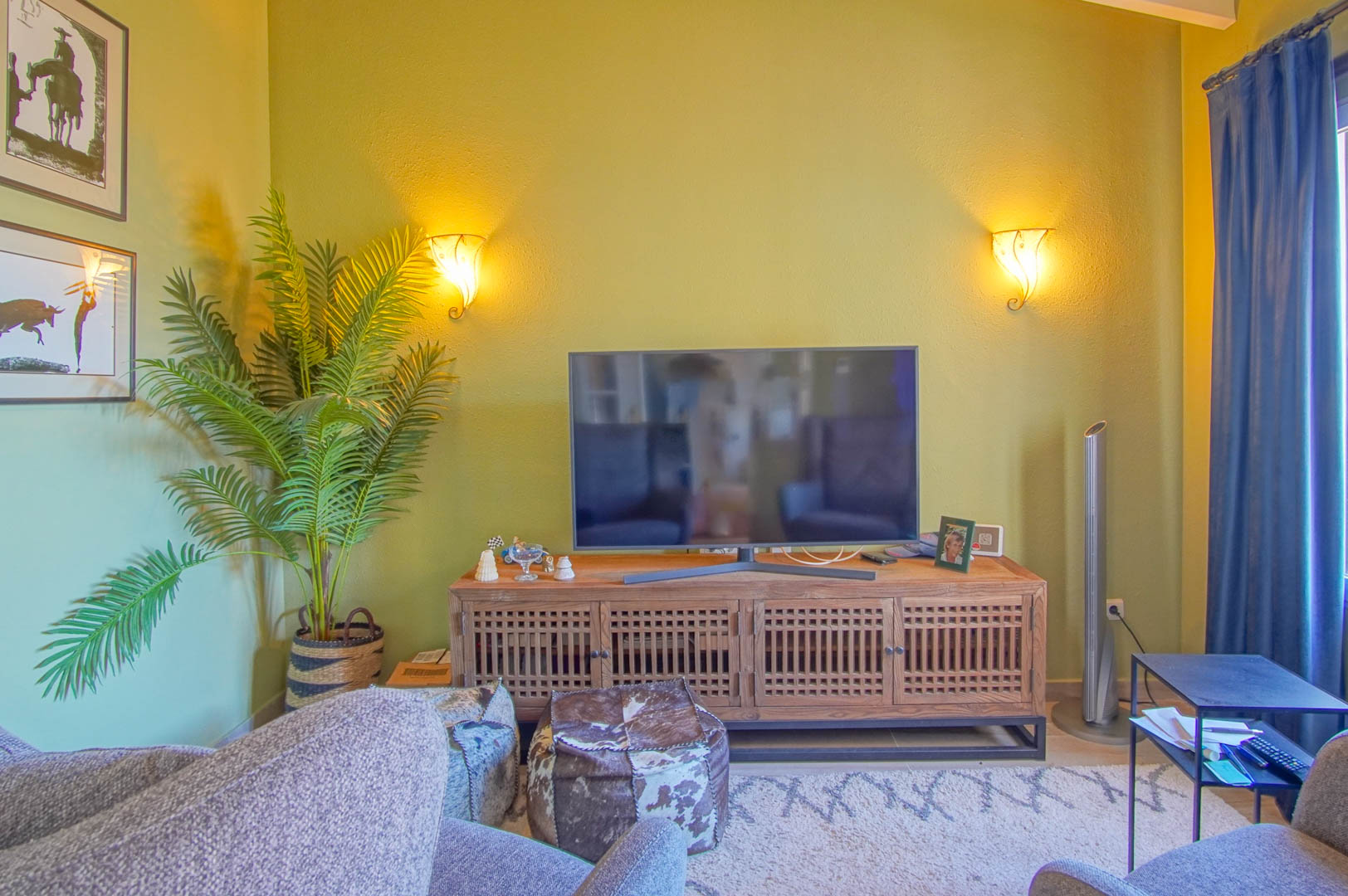 Zona de estar con mueble de televisión de madera tallada, iluminación cálida mediante apliques de pared y decoración con plantas naturales y pufs de piel.