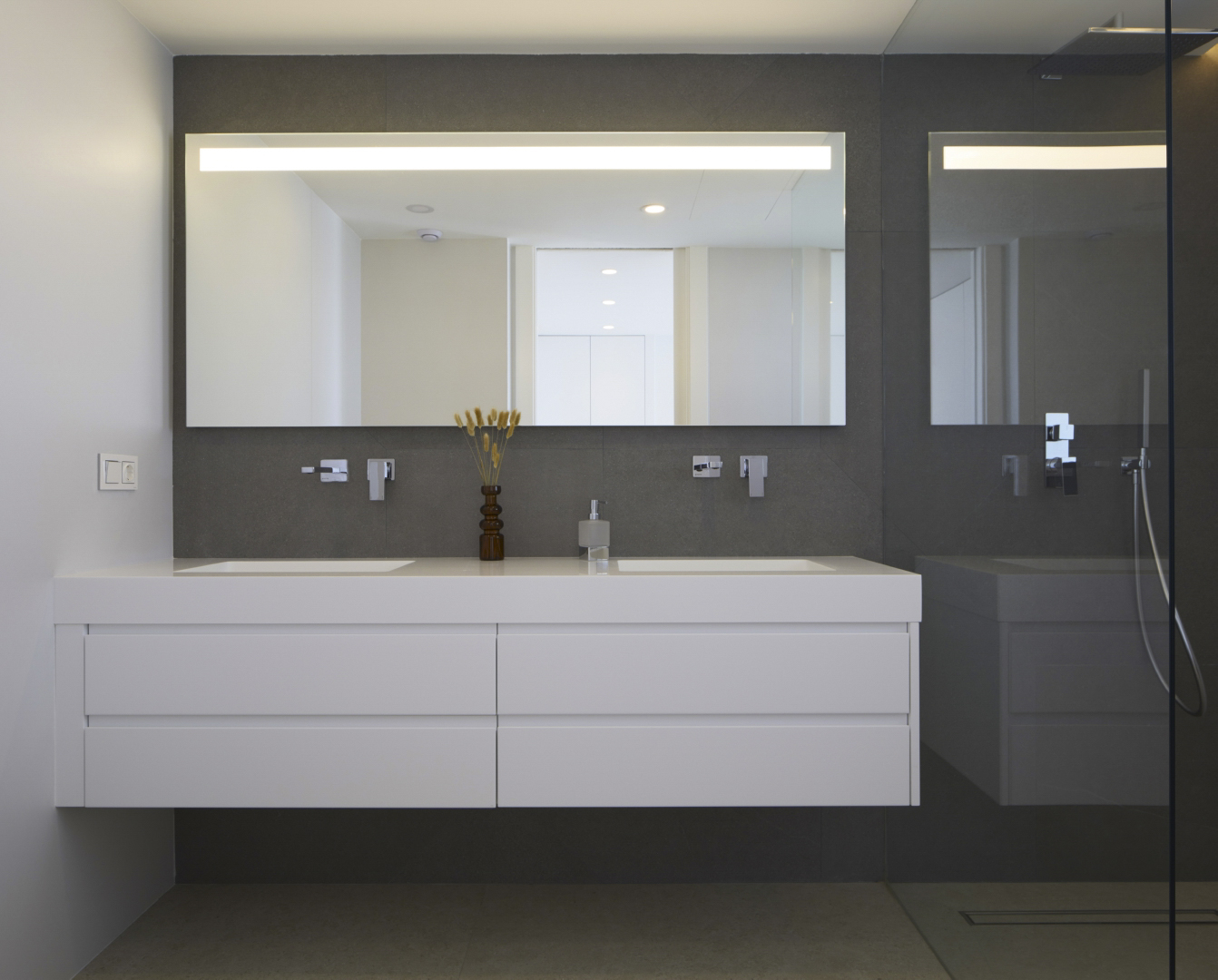 Baño moderno con mueble suspendido blanco de doble lavabo, grifería empotrada cromada y espejo retroiluminado sobre pared de gres porcelánico oscuro.