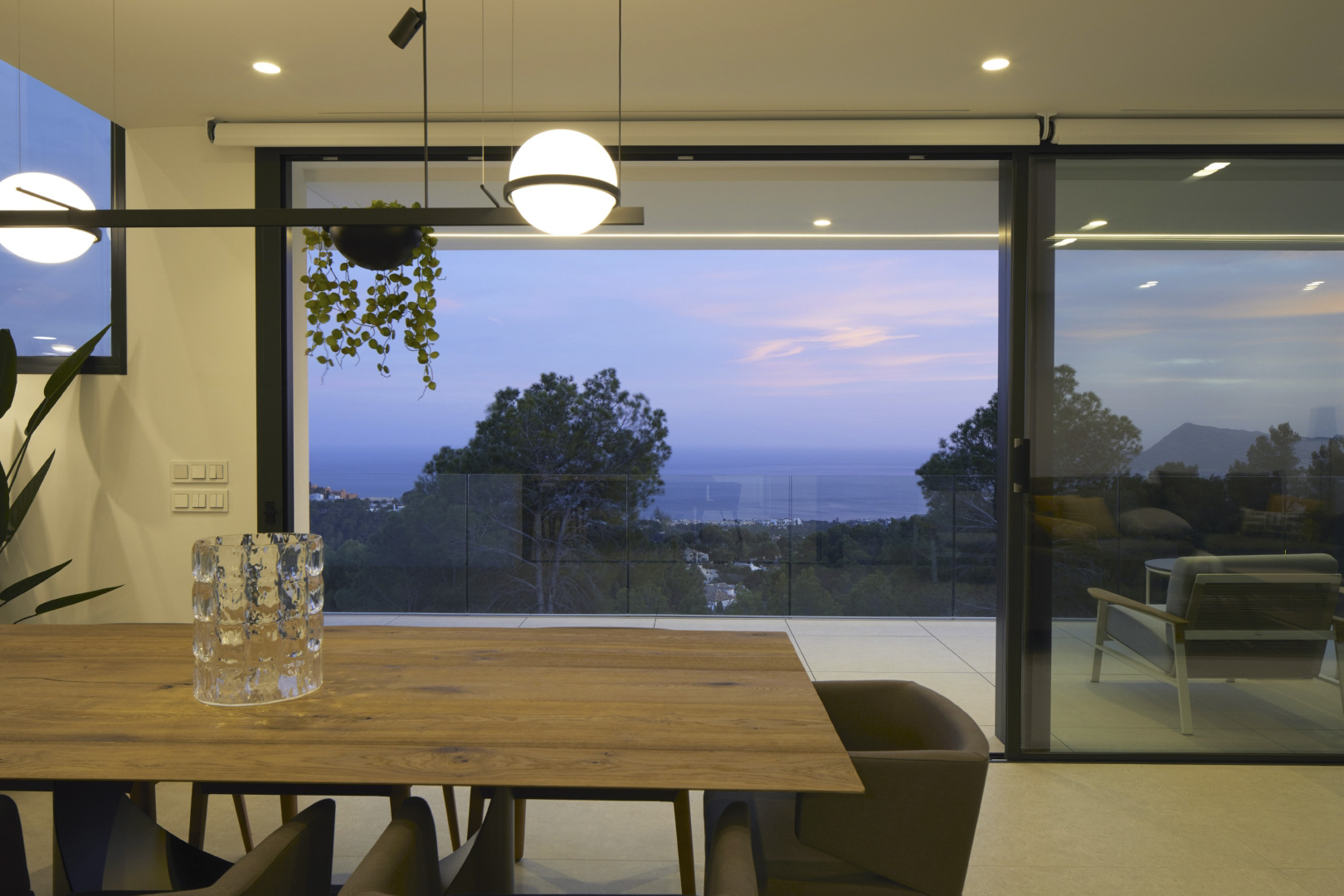 Comedor con mesa de madera natural y lámparas de diseño esféricas, con acceso directo a la terraza y vistas panorámicas al mar Mediterráneo al atardecer.