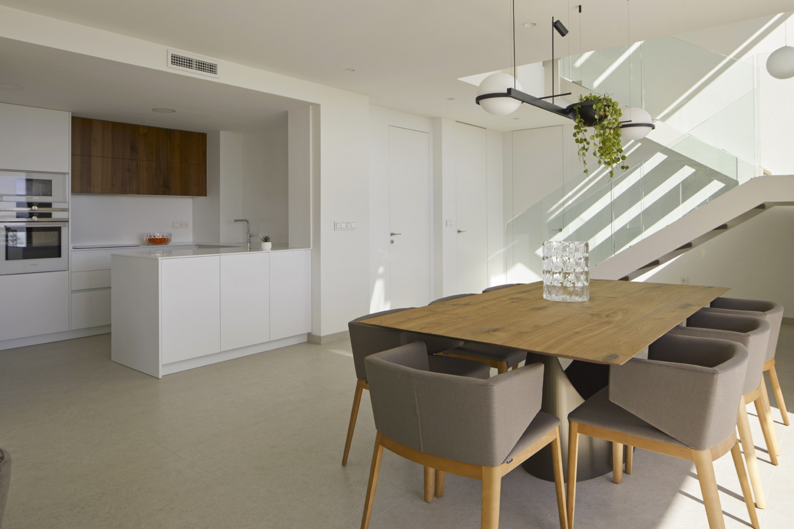 Comedor diáfano con mesa de madera y cocina integrada de diseño minimalista, destacando la escalera con barandilla de cristal y luz natural.
