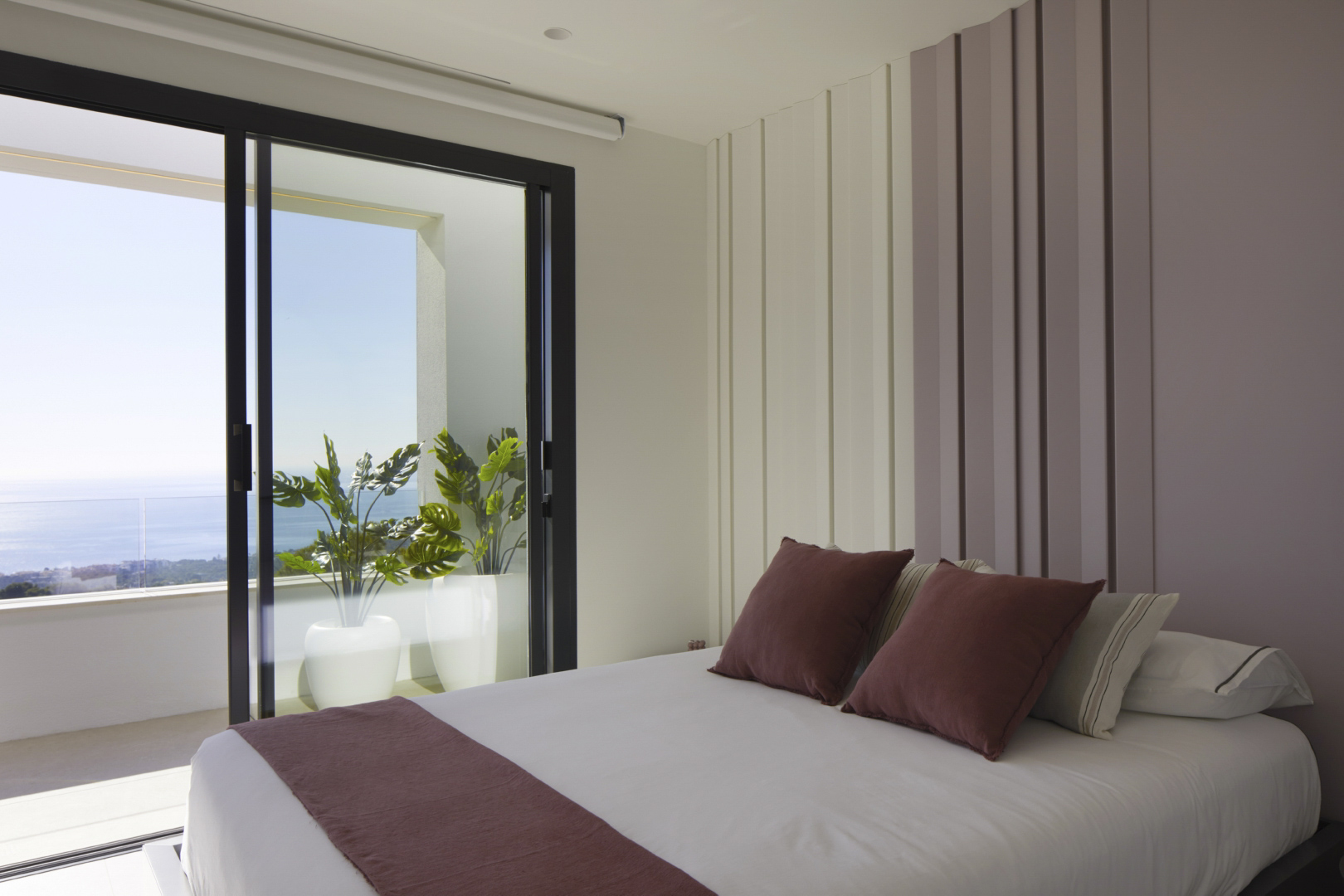Dormitorio con panelado vertical en tonos neutros y malva, con acceso directo a una terraza privada con plantas y vistas panorámicas al Mediterráneo.