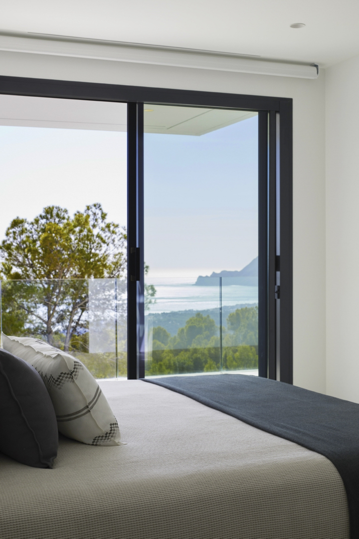 Dormitorio principal con ventanal de suelo a techo, carpintería oscura y vistas panorámicas al mar Mediterráneo y la Sierra de Altea.