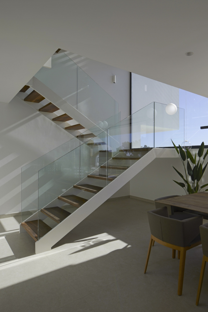 Escalera de diseño con peldaños de madera oscura y barandillas de cristal, integrada en un espacio diáfano con luz natural y estructura metálica blanca.