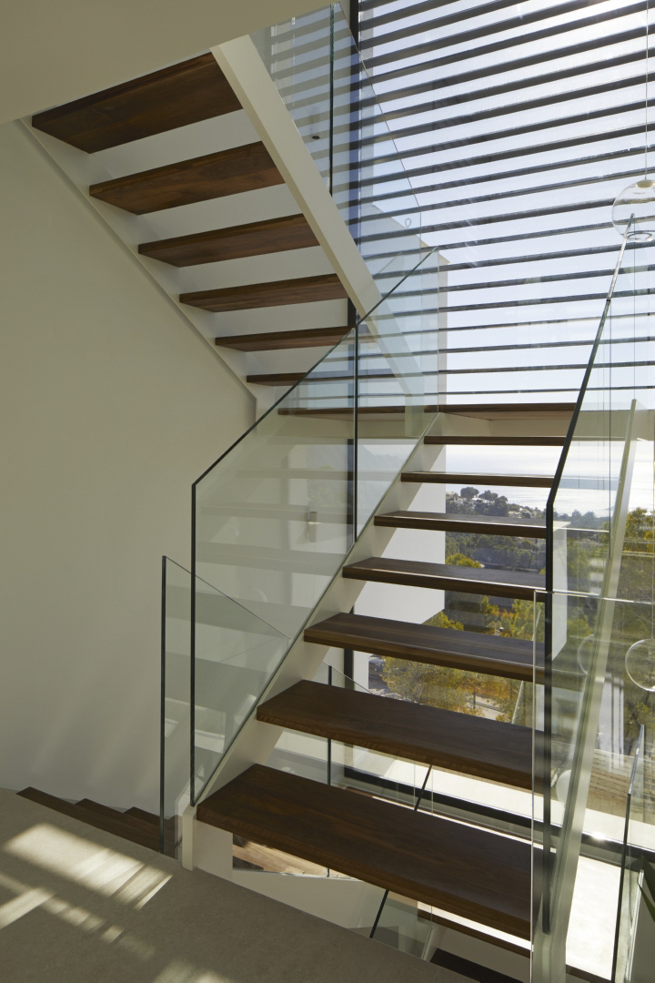 Escalera de diseño con peldaños de madera oscura y barandillas de cristal, situada frente a un gran ventanal con vistas panorámicas al mar Mediterráneo.