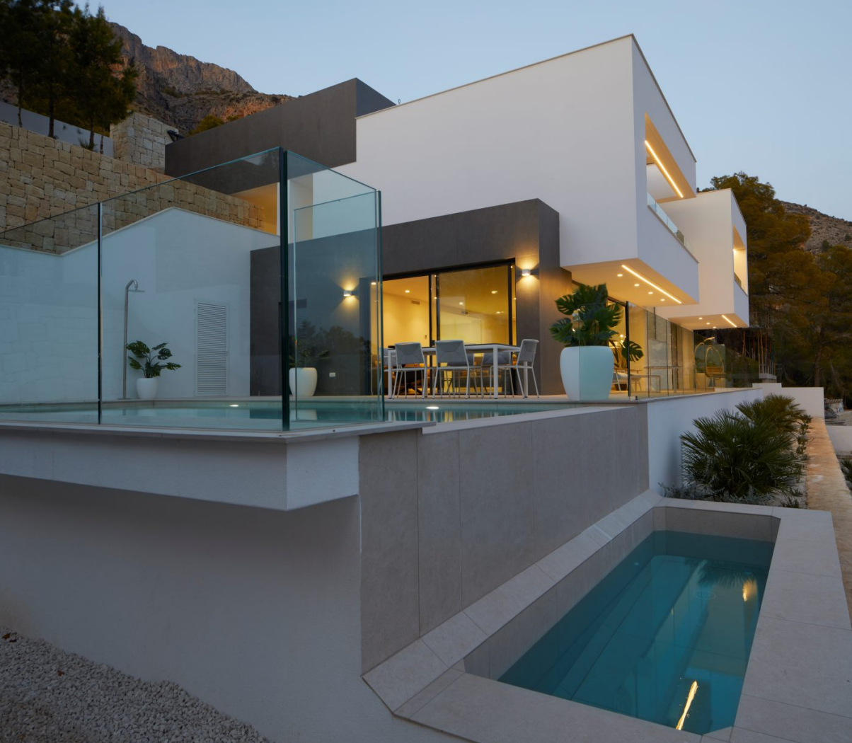 Fachada exterior de diseño contemporáneo con piscina infinita, barandillas de cristal y terrazas iluminadas integradas en la ladera de la Sierra de Altea.