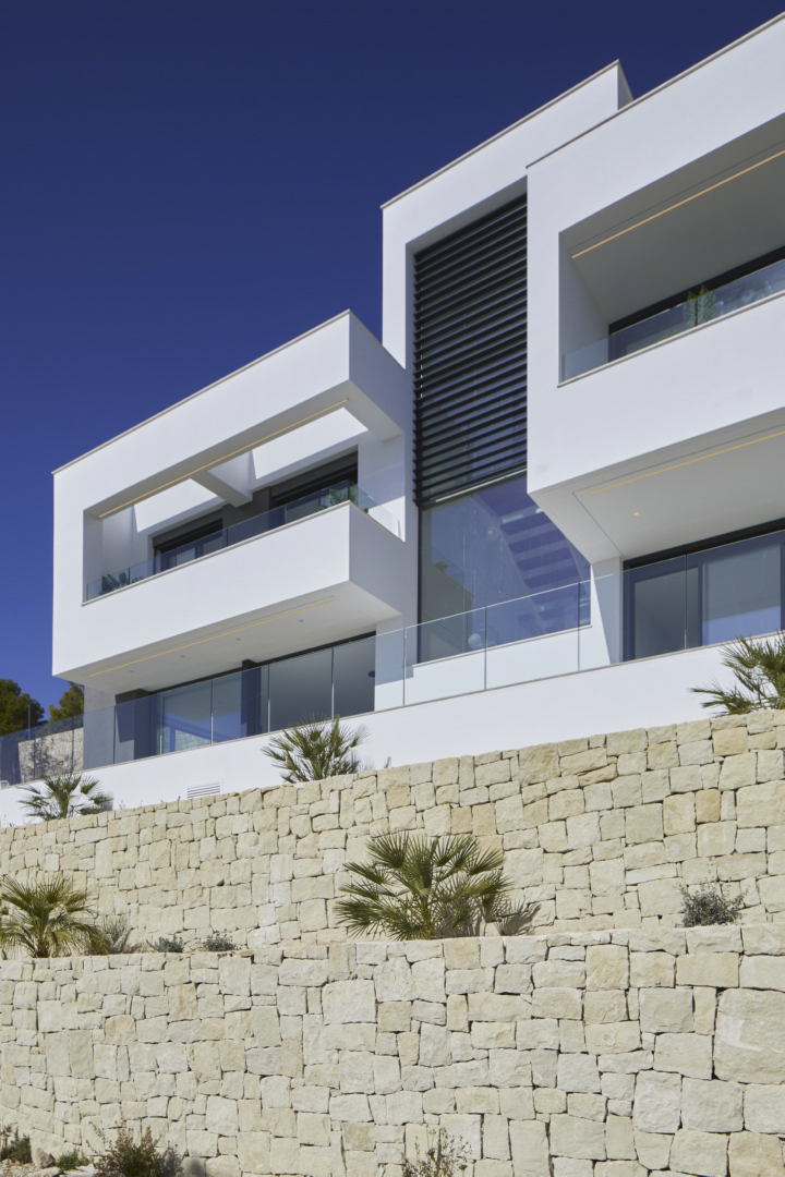 Fachada moderna de líneas geométricas blancas con barandillas de cristal, situada sobre un muro de piedra seca natural con vegetación mediterránea.