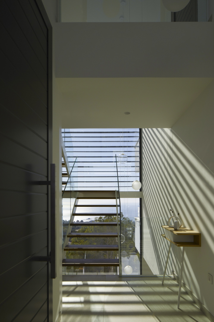 Hall de entrada con escalera de peldaños de madera y barandilla de cristal. Destaca la iluminación natural con sombras lineales y vistas al mar a través del gran ventanal.