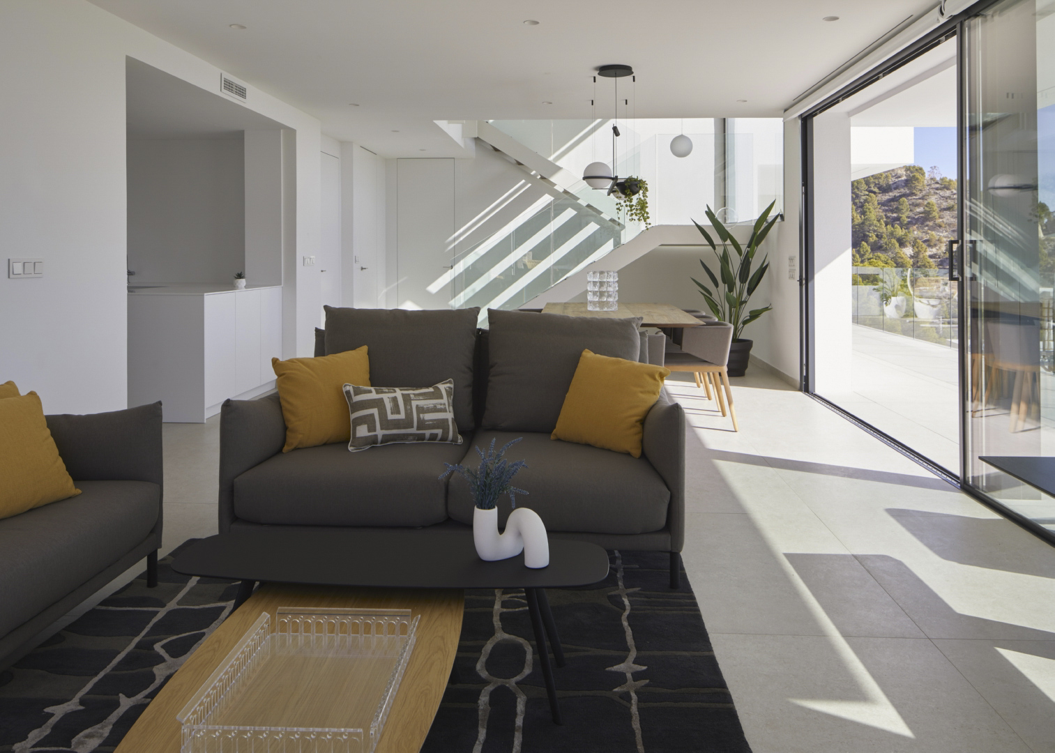 Salón comedor de planta abierta con sofás grises, cojines mostaza y grandes ventanales correderos que integran la terraza y la luz natural.