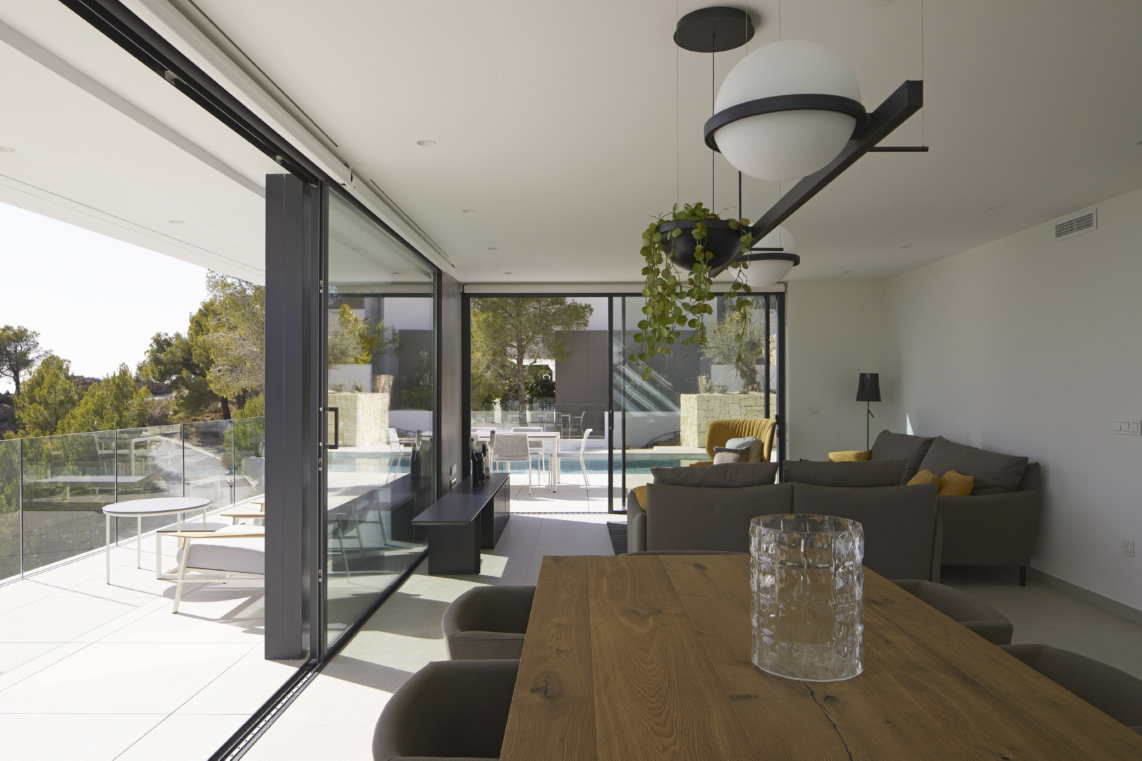 Salón-comedor diáfano con mesa de madera maciza, grandes ventanales correderos que conectan con la terraza y vistas a la piscina infinity y el entorno natural.